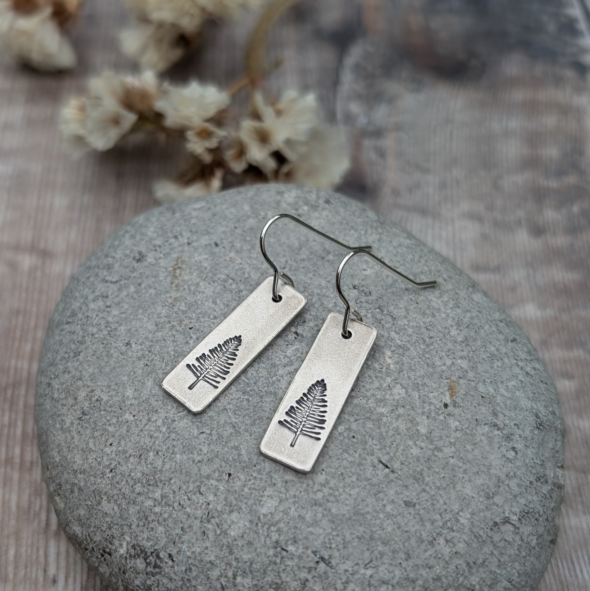 A pair of oxidised silver rectangle earrings featuring an etched evergreen tree design, displayed on a smooth grey stone. Softly blurred dried flowers in the background enhance the natural theme.