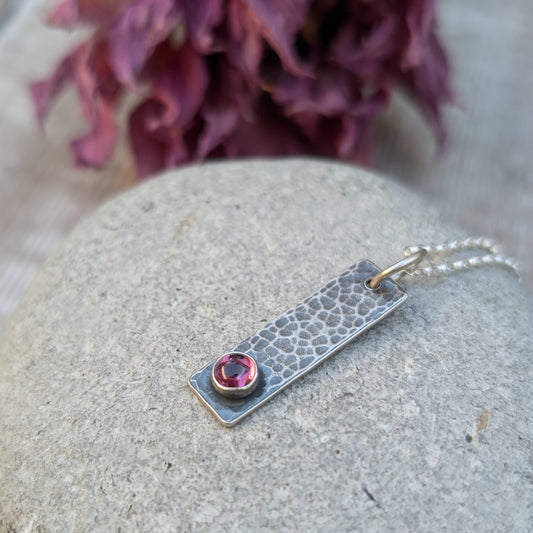 Pendant is an oxidised Sterling Silver rectangle with hammered finish, suspended from a silver chain via silver open circle link. Set onto the bottom of the rectangle is a small, round Rhodolite gemstone, dark pink in colour.