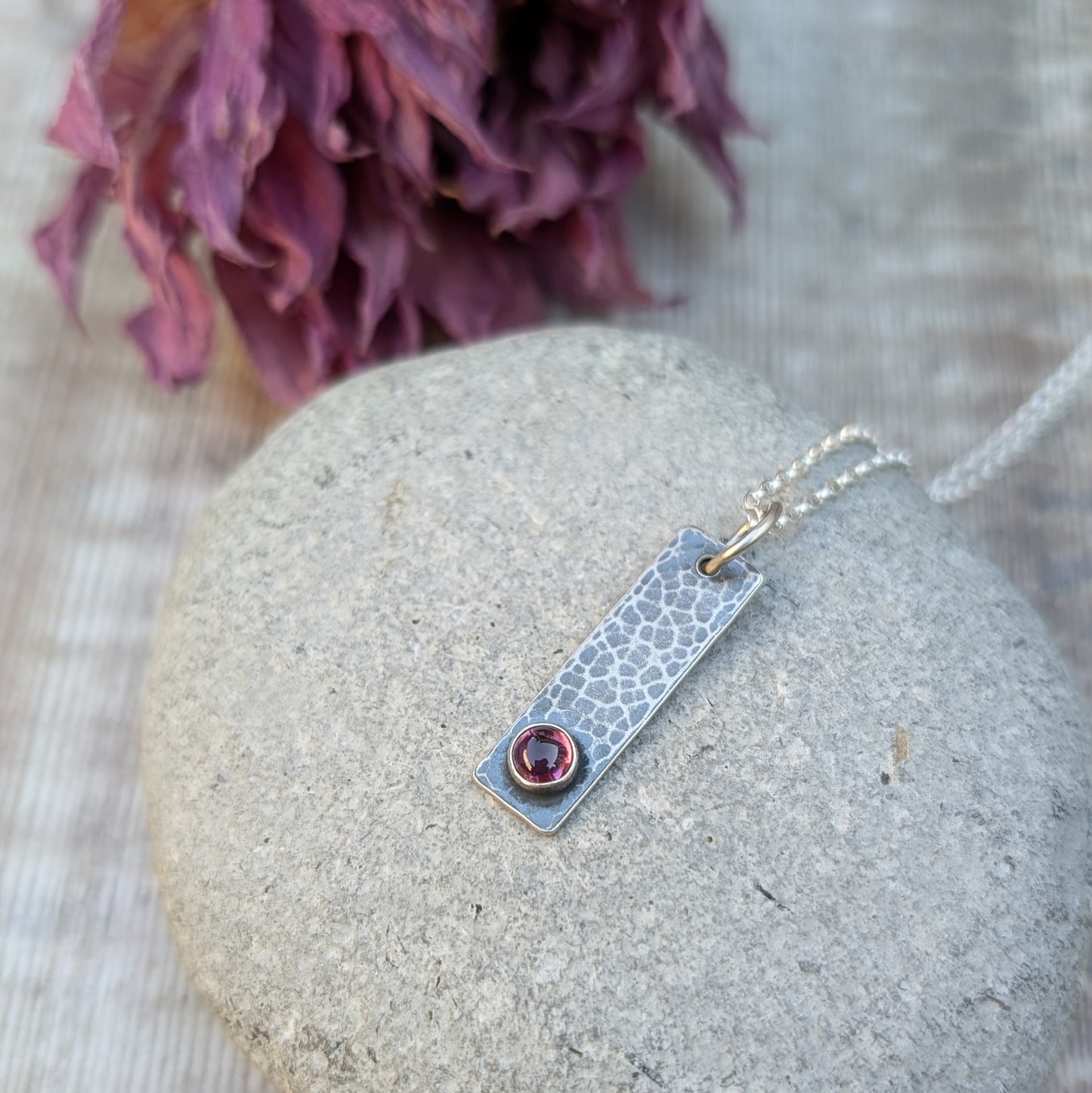Pendant is an oxidised Sterling Silver rectangle with hammered finish, suspended from a silver chain via silver open circle link. Set onto the bottom of the rectangle is a small, round Rhodolite gemstone, dark pink in colour.