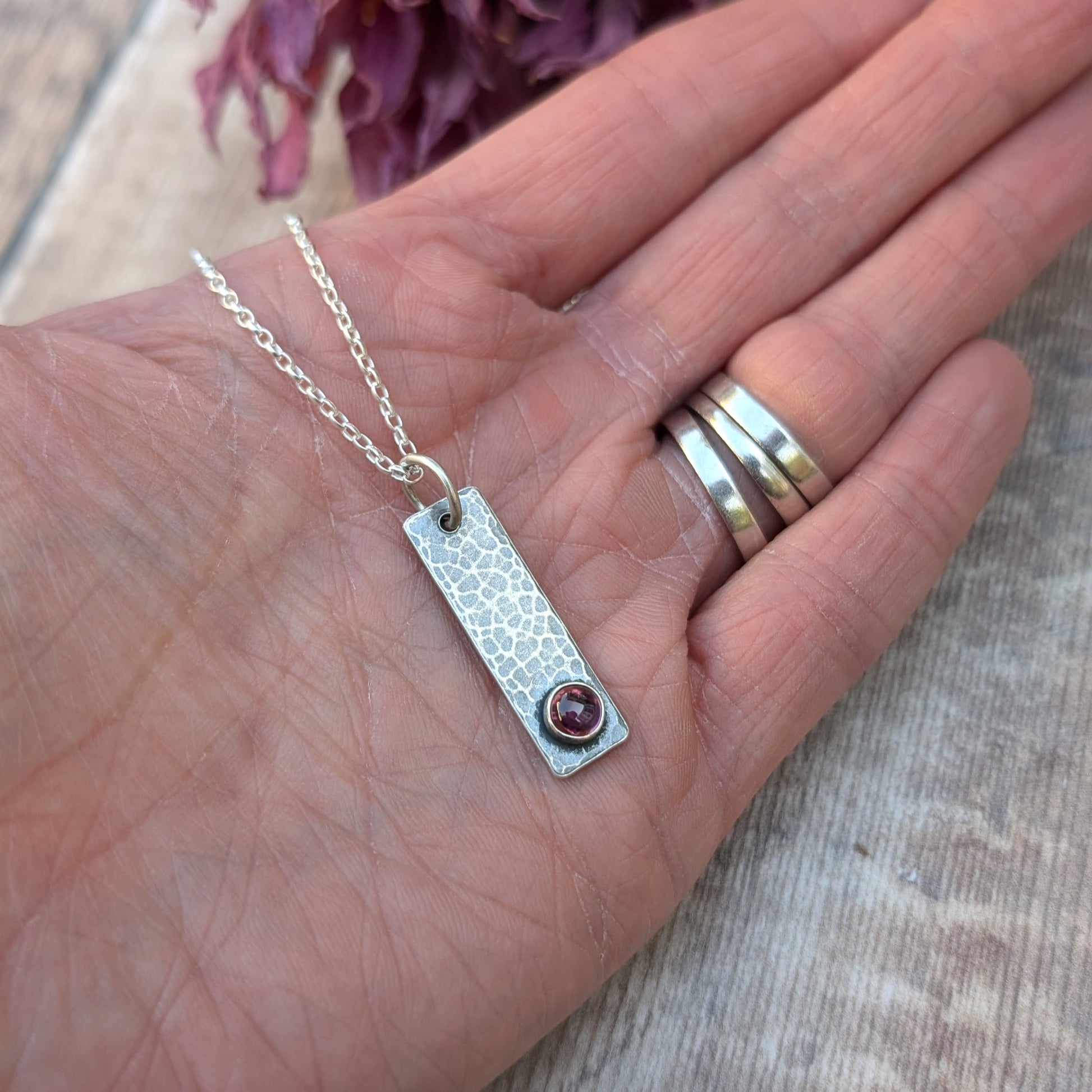 Pendant is an oxidised Sterling Silver rectangle with hammered finish, suspended from a silver chain via silver open circle link. Set onto the bottom of the rectangle is a small, round Rhodolite gemstone, dark pink in colour.