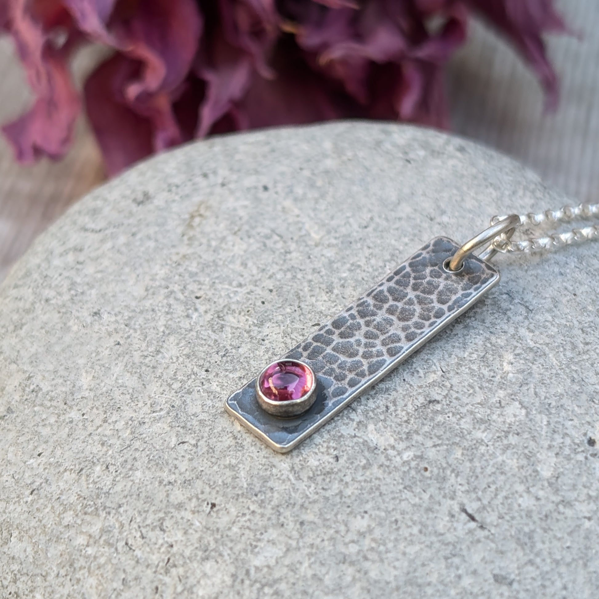 Pendant is an oxidised Sterling Silver rectangle with hammered finish, suspended from a silver chain via silver open circle link. Set onto the bottom of the rectangle is a small, round Rhodolite gemstone, dark pink in colour.