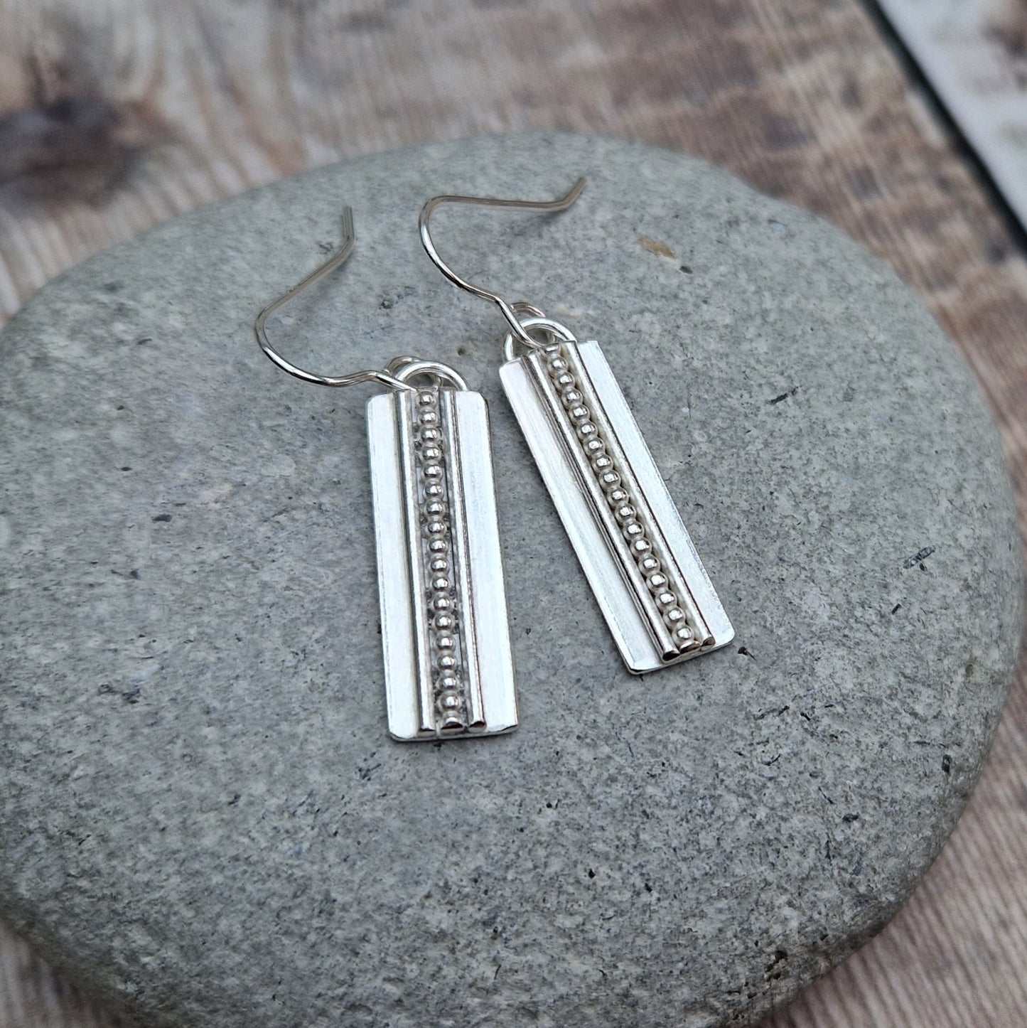Each earring is a Sterling Silver rectangle suspended from silver earring wire. Along the centre of the rectangle is a raised beaded line with a raised smooth line on each side, running top to bottom of the rectangle.