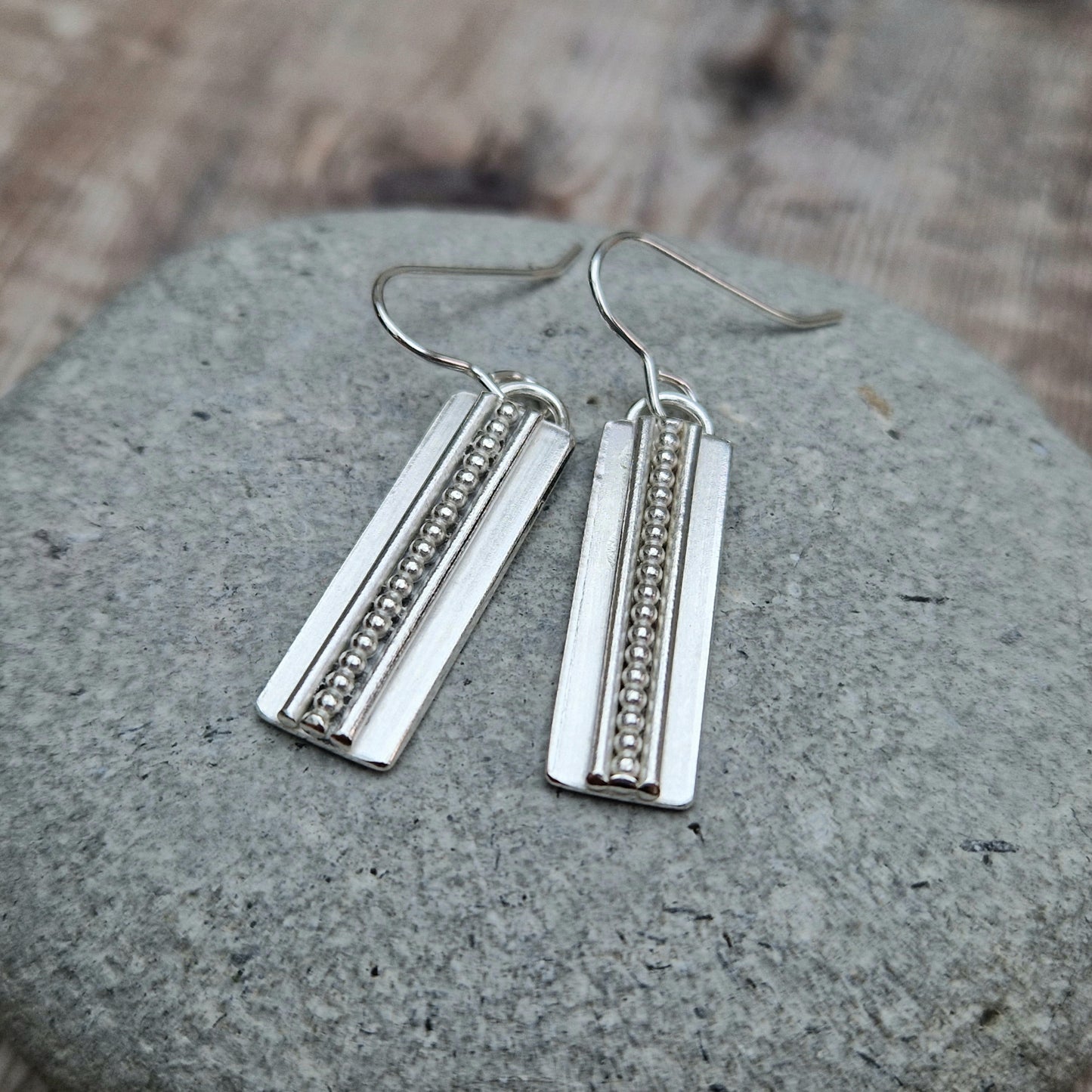 Each earring is a Sterling Silver rectangle suspended from silver earring wire. Along the centre of the rectangle is a raised beaded line with a raised smooth line on each side, running top to bottom of the rectangle.