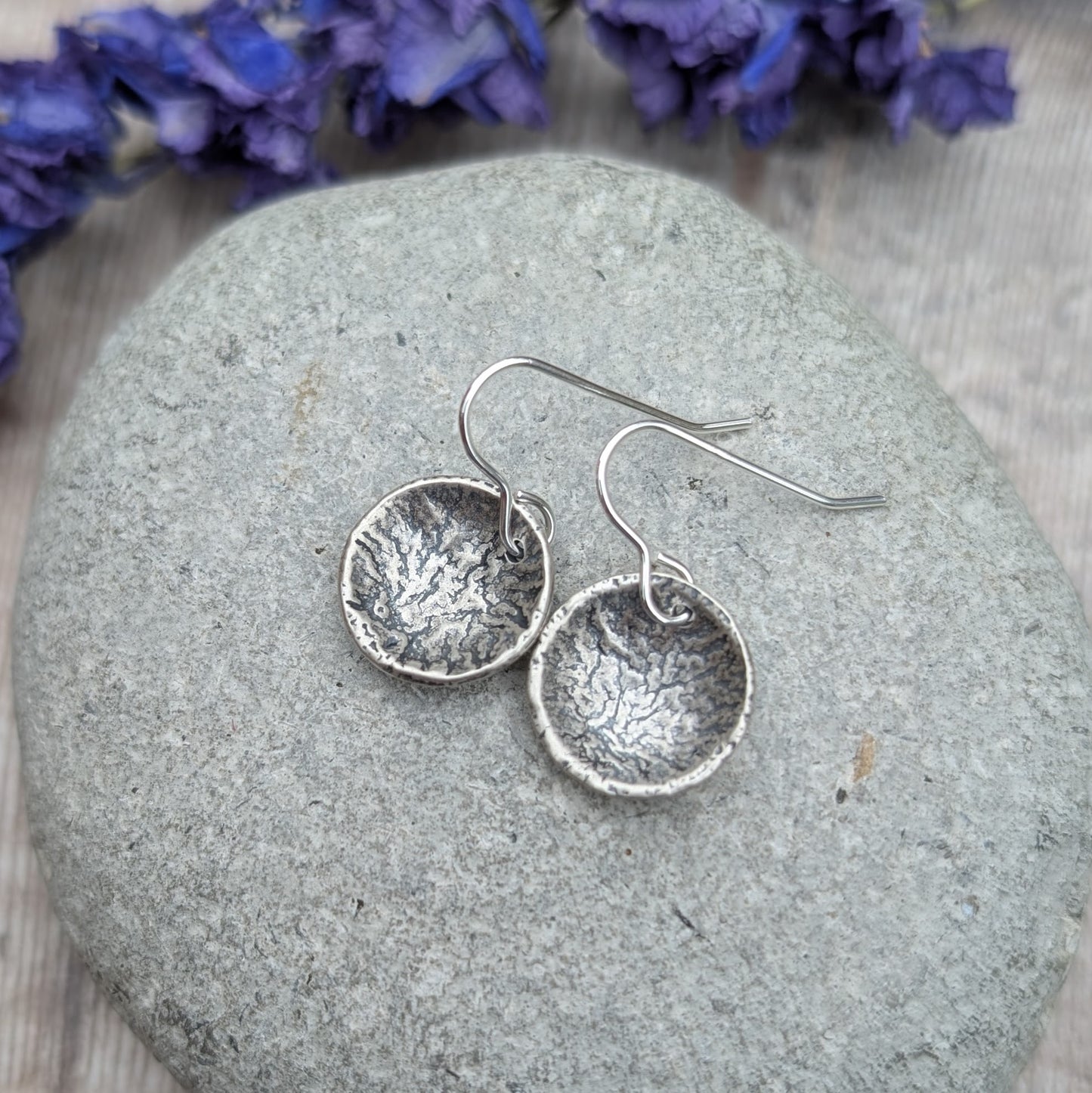 Pair of oxidised reticulated silver disc earrings resting on a grey stone with purple flowers in background.