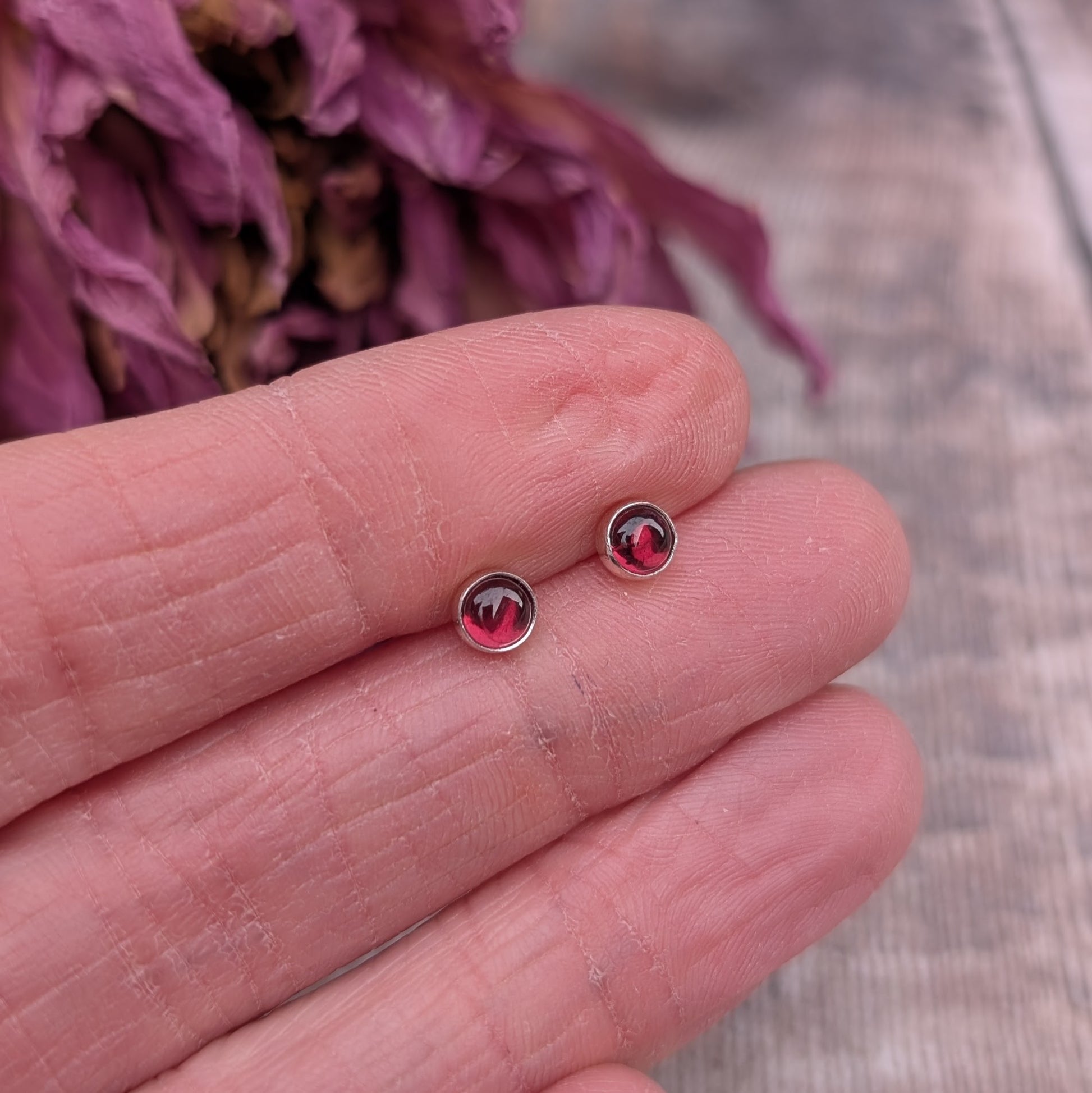 A close-up of a pair of small rhodolite garnet stud earrings set in recycled sterling silver, resting on a person's fingers. The deep red gemstones catch the light against a backdrop of dried purple flowers and a rustic wooden surface.