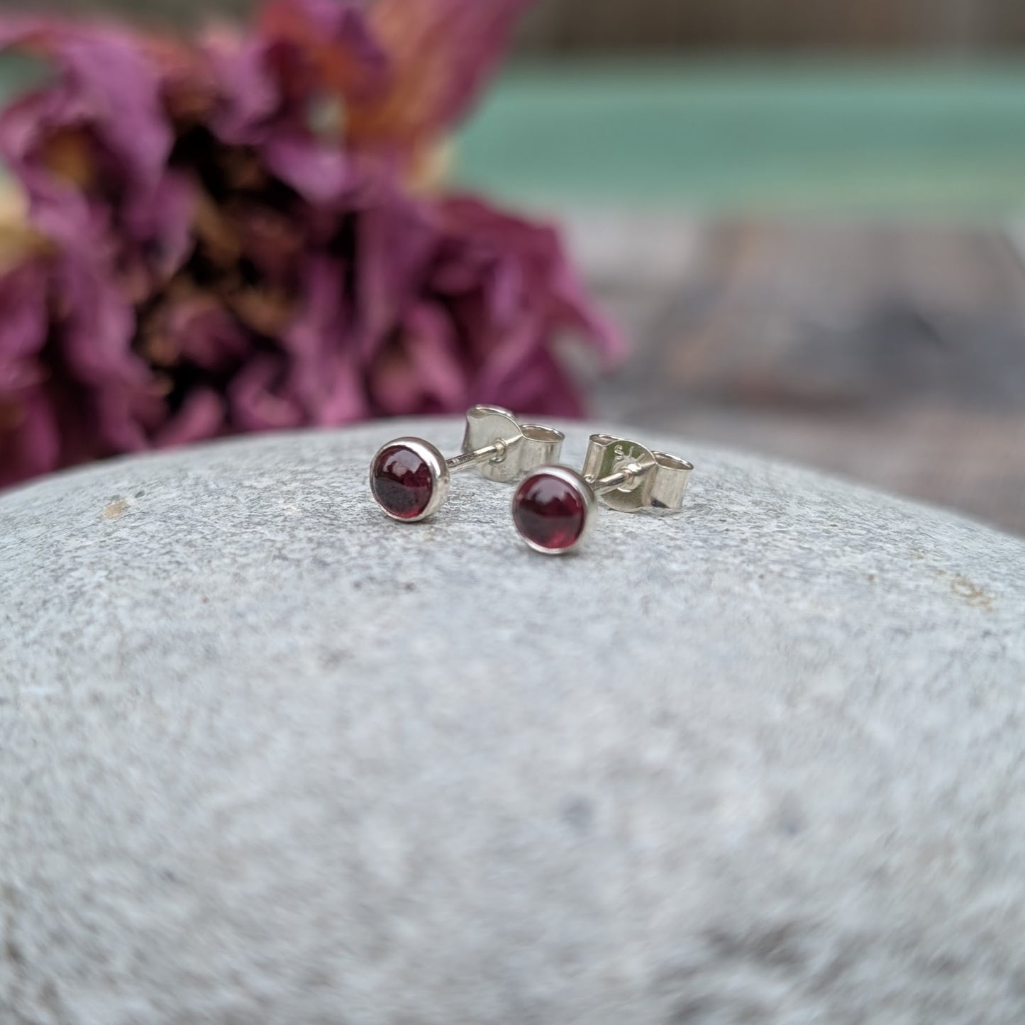 A pair of rhodolite garnet stud earrings displayed on a smooth, light grey stone. The silver setting enhances the deep red hues of the garnet, with a softly blurred background featuring dried purple flowers.