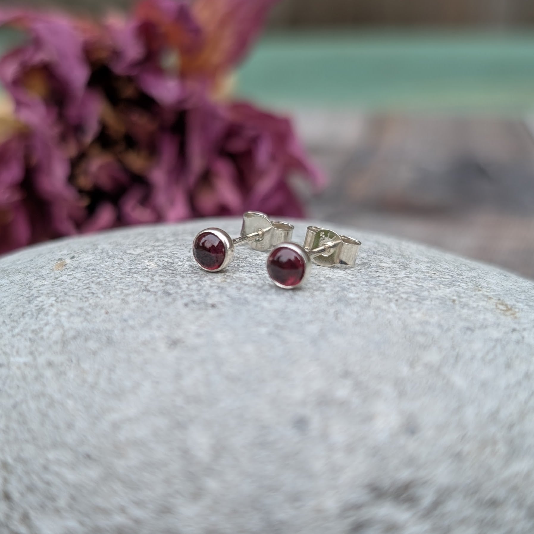 A pair of rhodolite garnet stud earrings displayed on a smooth, light grey stone. The silver setting enhances the deep red hues of the garnet, with a softly blurred background featuring dried purple flowers.