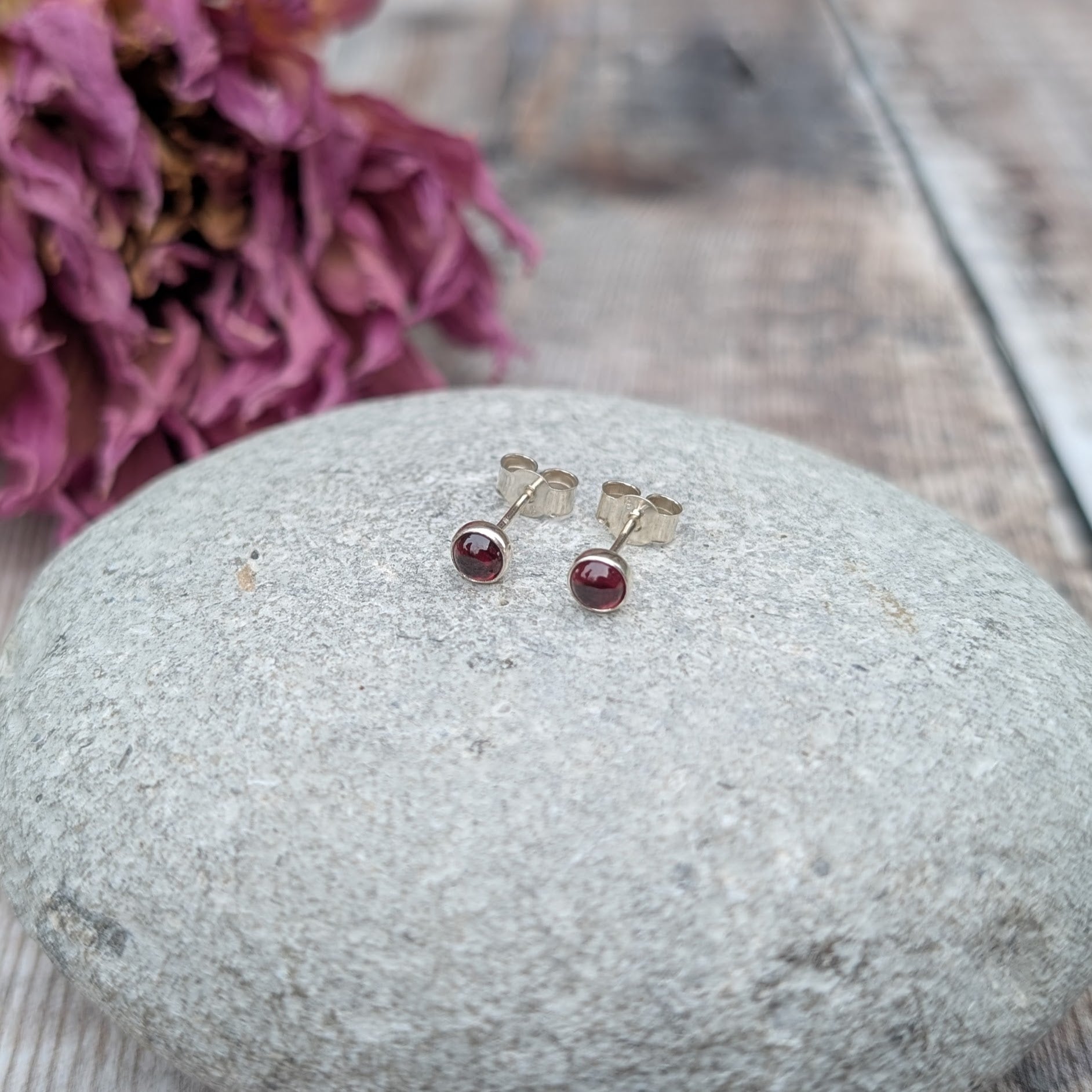 A pair of rhodolite garnet stud earrings displayed on a smooth, light grey stone. The silver setting enhances the deep red hues of the garnet, with a softly blurred background featuring dried purple flowers.