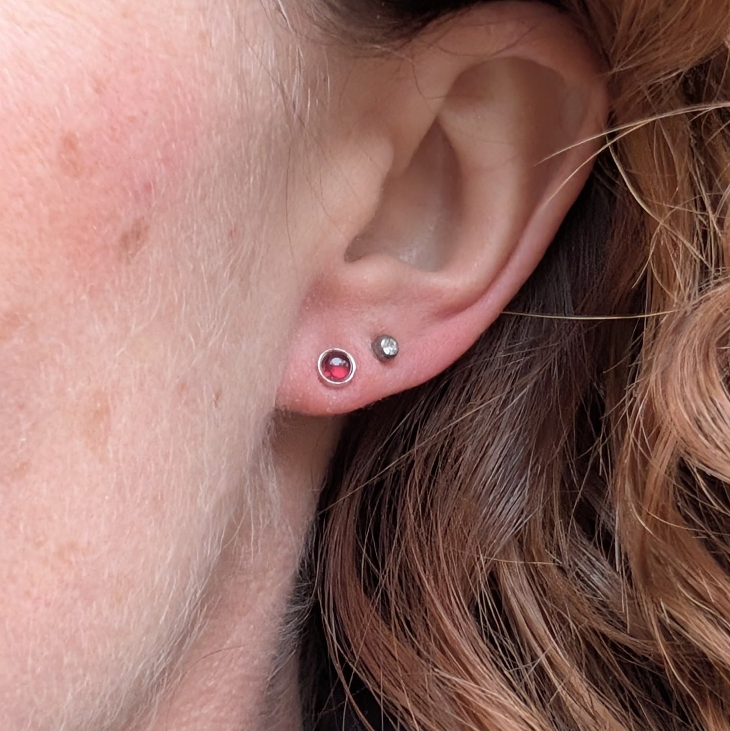 A close-up of a person wearing one of the rhodolite garnet stud earrings in their ear. The garnet's deep red color contrasts with their skin and hair, with a smaller silver stud earring worn alongside it.