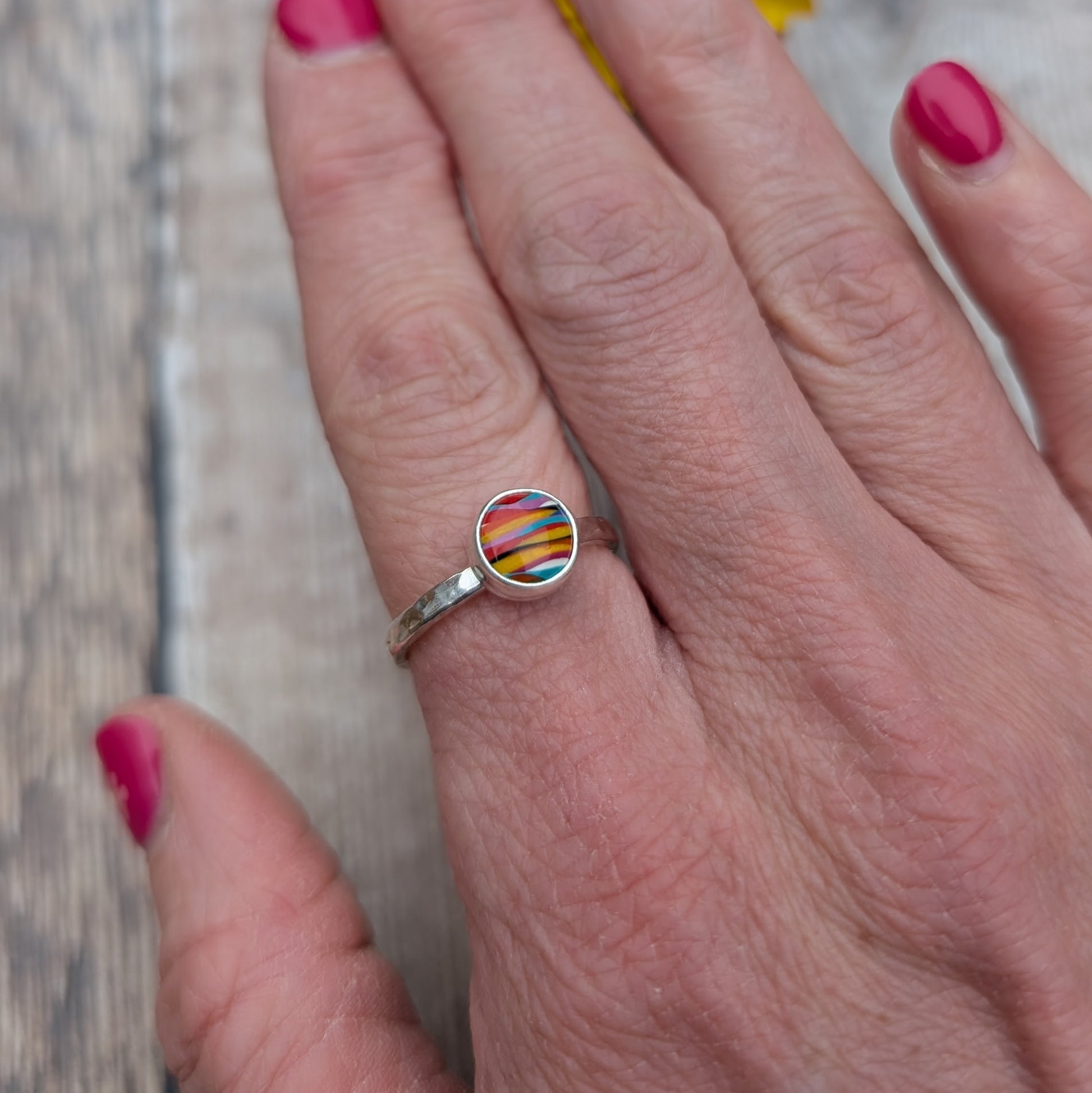 Close-up of a colourful round Surfite ring with a hammered sterling silver band, worn on a hand with pink nail polish.