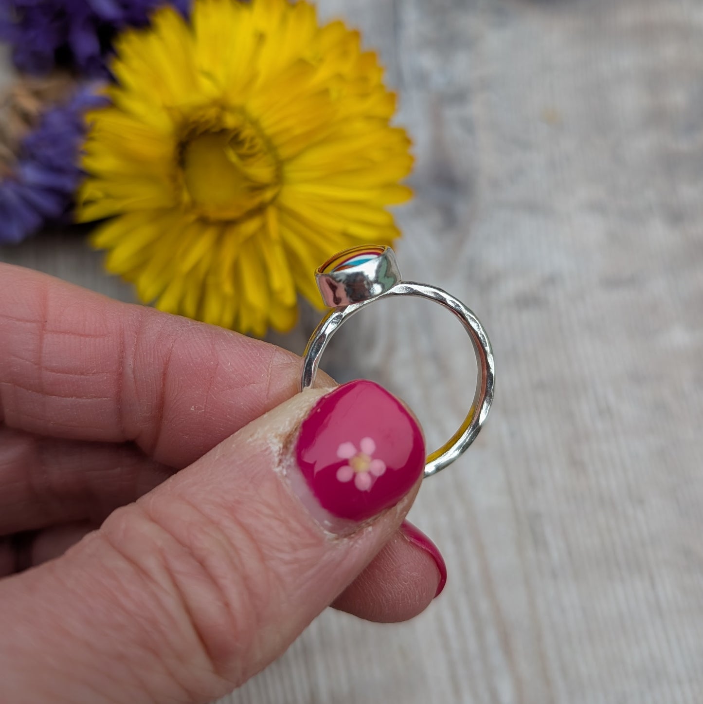 Side view of the Surfite ring showing the bezel setting and hammered silver band, held in front of a yellow flower.