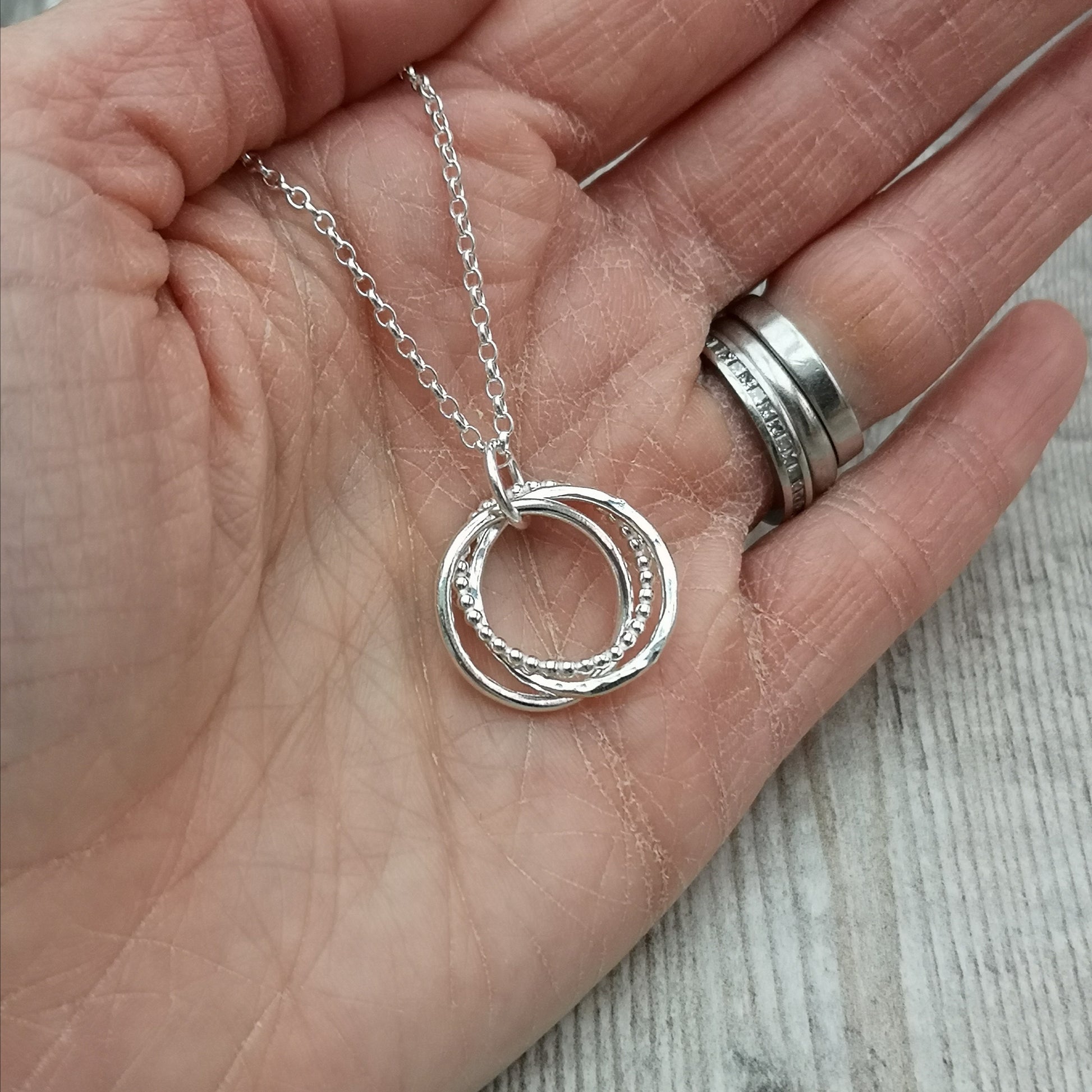 The silver necklace held in an open hand, showcasing the size and delicate details of the three textured, intertwined rings on the pendant.