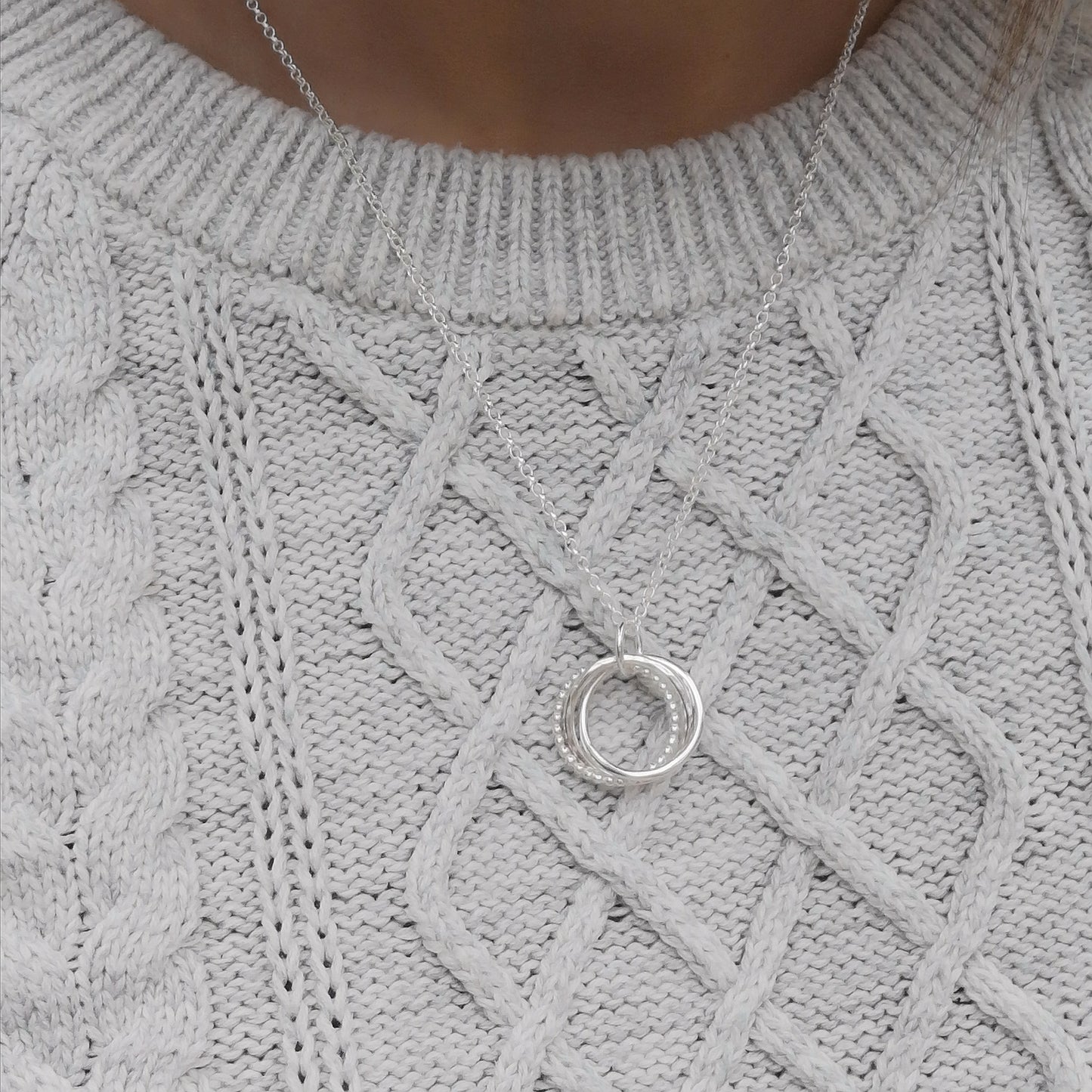 The silver necklace worn over a light grey cable-knit sweater, highlighting how the three-ring pendant sits on the chest, demonstrating the necklace's length and subtle elegance.