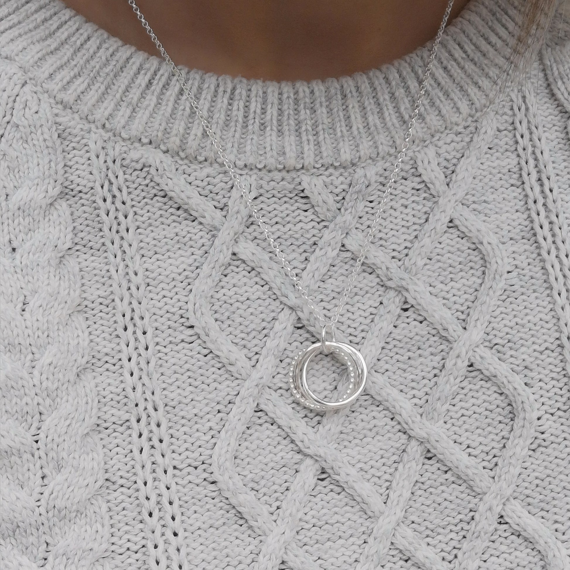 The silver necklace worn over a light grey cable-knit sweater, highlighting how the three-ring pendant sits on the chest, demonstrating the necklace's length and subtle elegance.