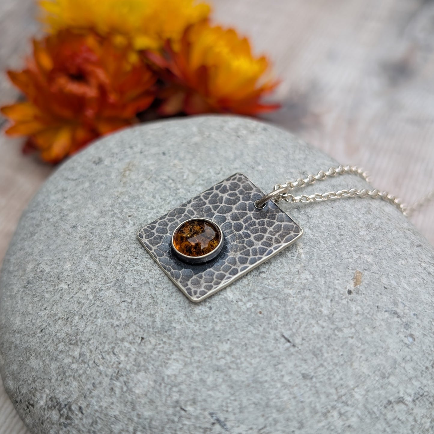 Close-up of the Silver Rectangle Necklace with Amber lying on a smooth grey stone, highlighting the hammered texture and deep golden tones of the amber.