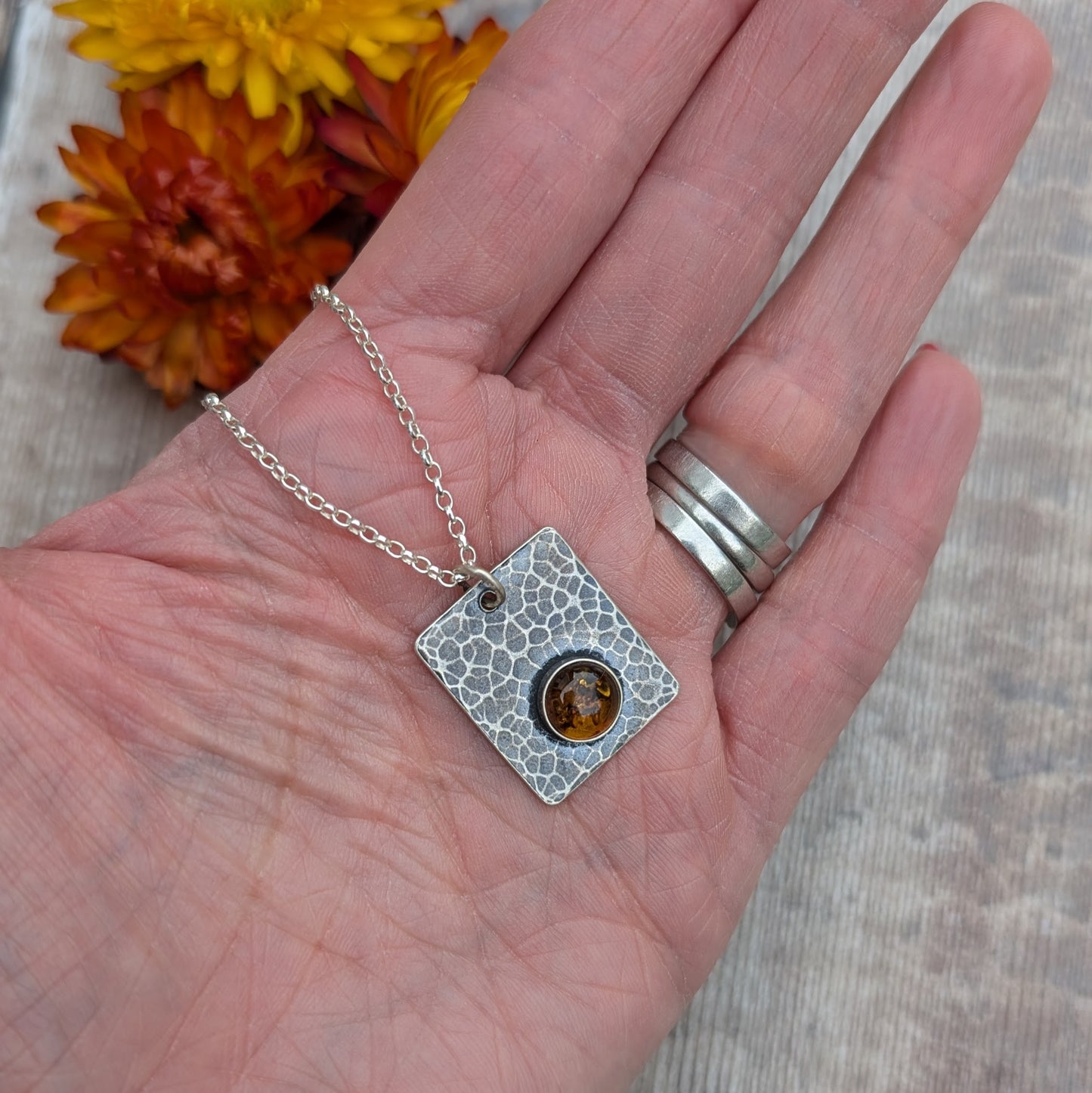 Another view of the Silver Rectangle Necklace with Amber in a hand, with orange flowers in the background.