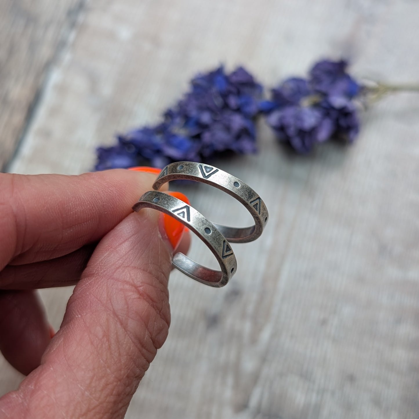 Close-up of a hand holding the silver geometric hoop earrings, with a backdrop of blurred purple flowers and wooden surface.