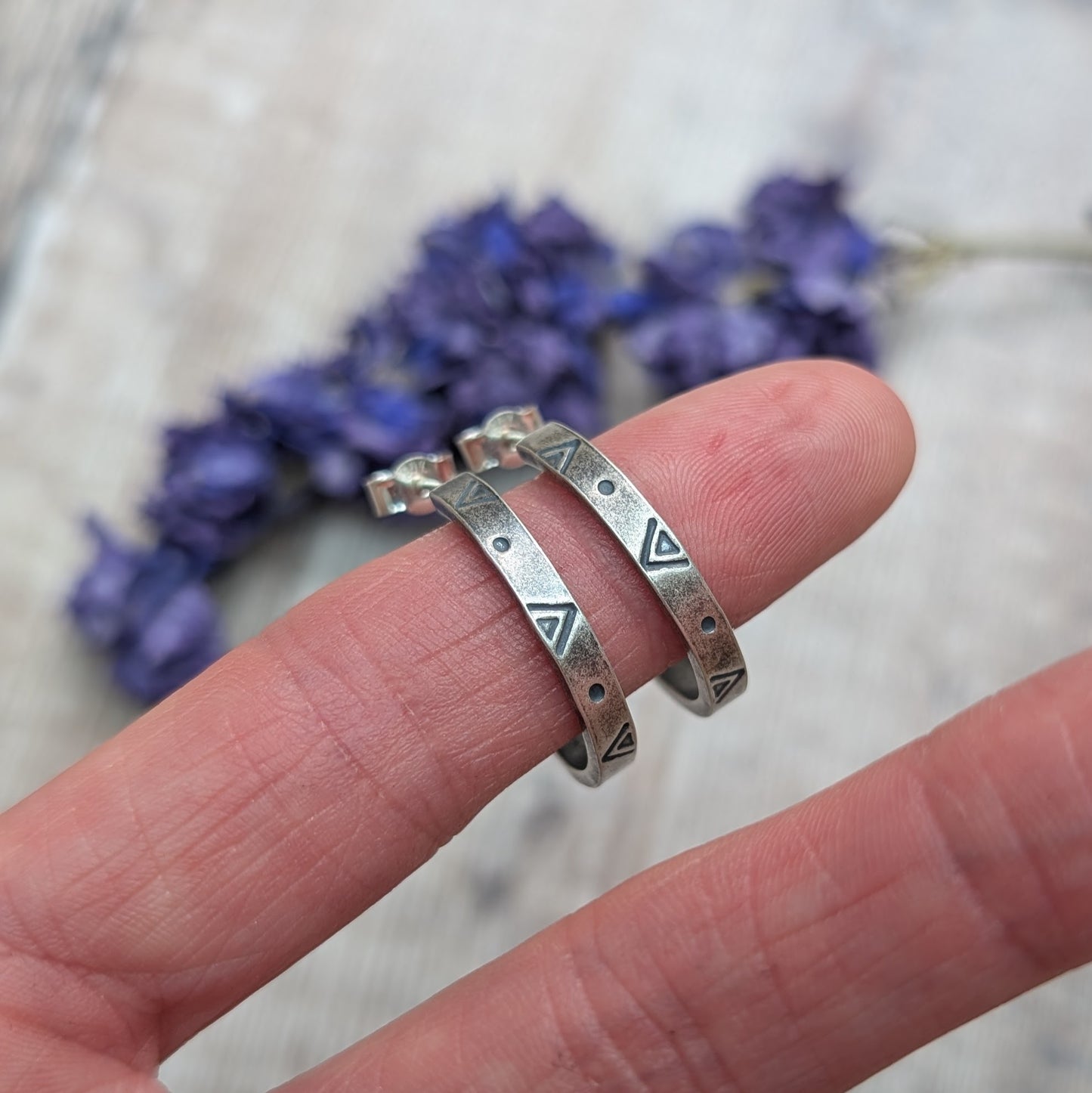 Silver geometric hoop earrings balanced across a finger, showing their hand-stamped triangle and dot pattern.