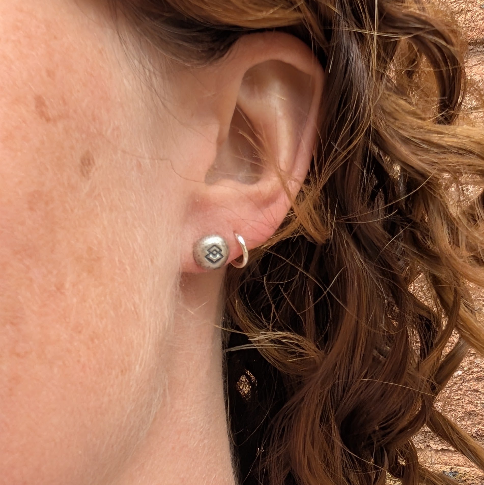 The Silver Geometric Pebble Studs being worn in the lobe of an ear, paired with a small silver hoop in the second piercing
