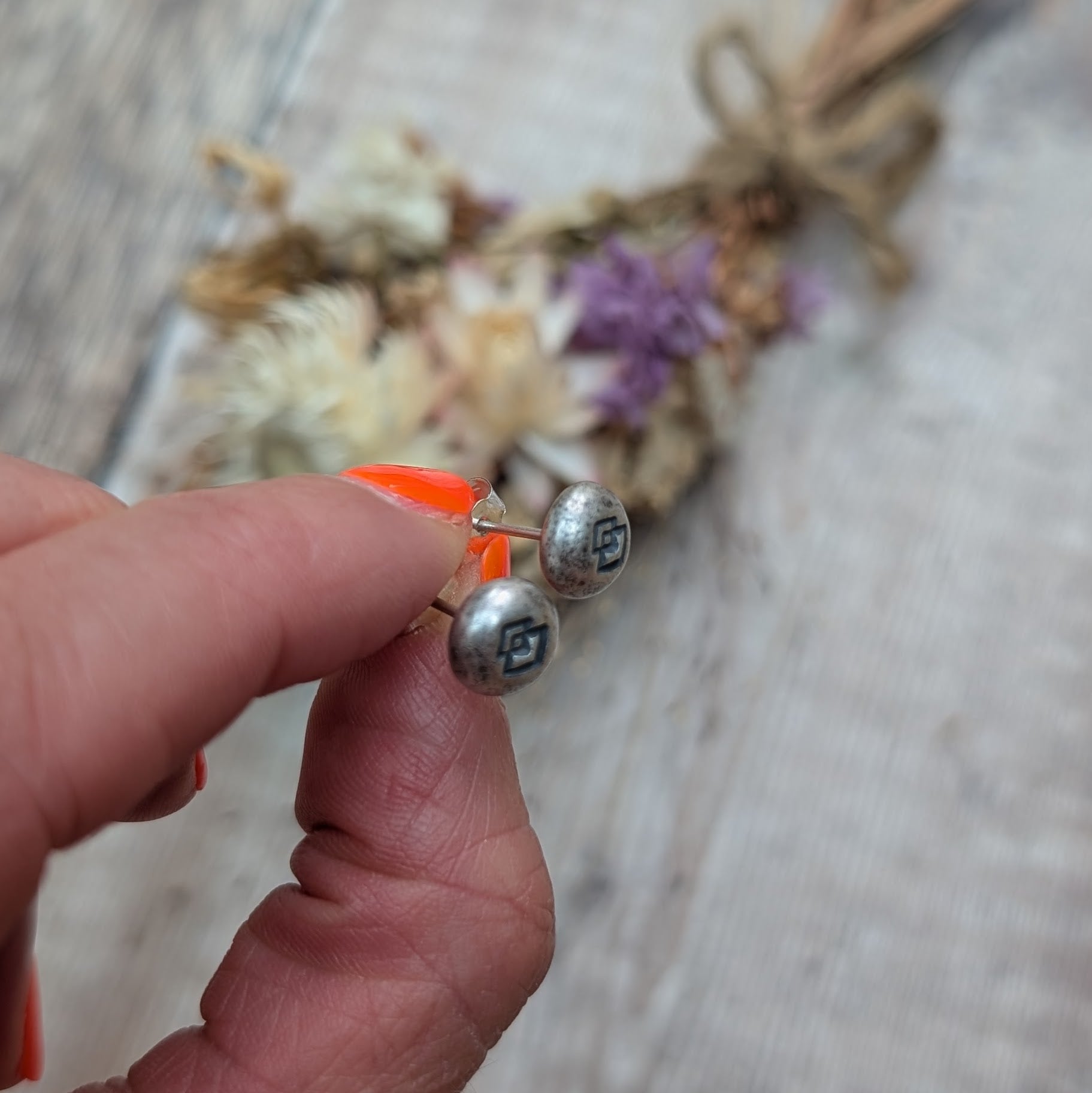 A close-up of the Silver Geometric Pebble Studs being held between two fingers, with a rustic wood and dried flower background.