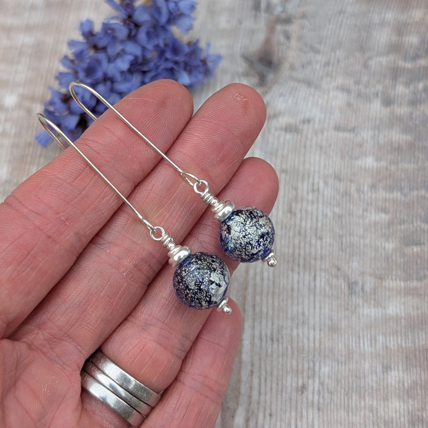 Silver blue venetian glass earrings displayed on a hand with dried flowers in the background.