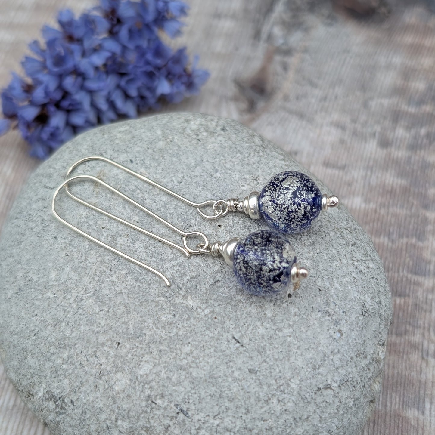 Silver blue venetian glass earrings displayed on a grey stone with dried flowers in the background.