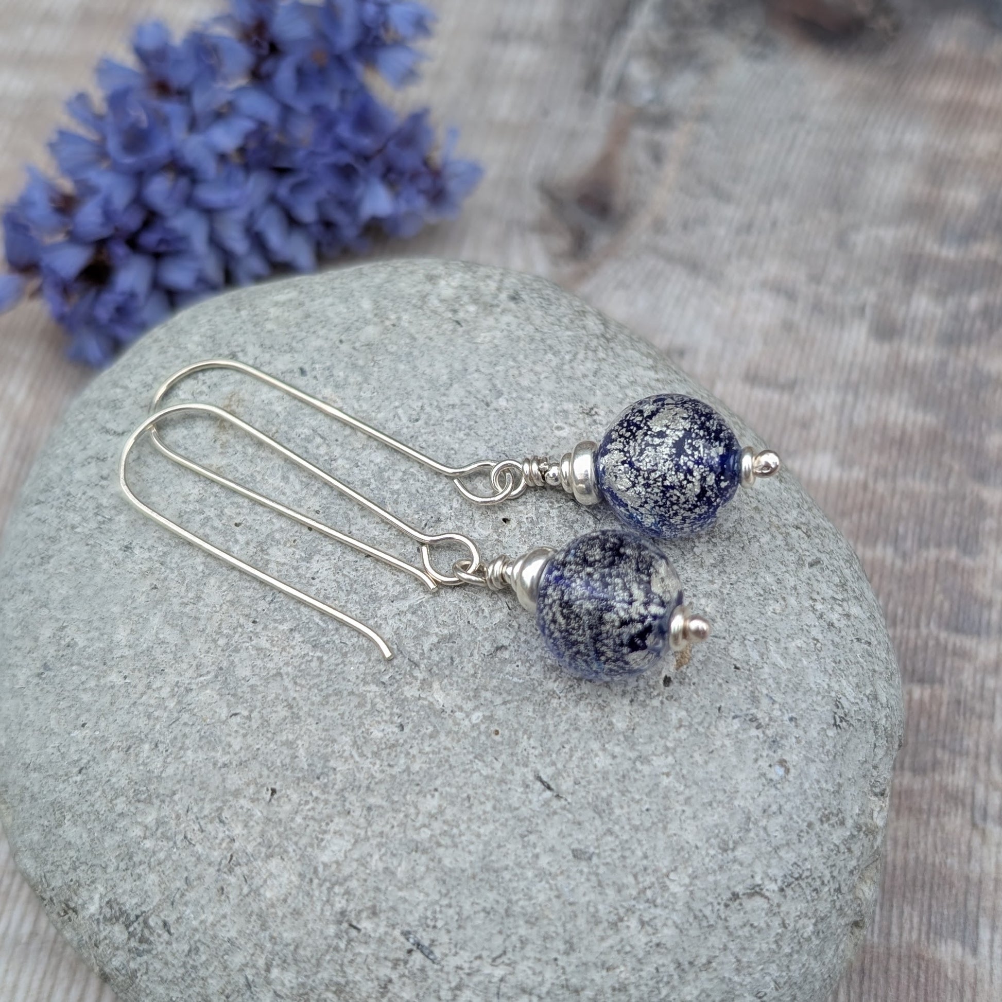 Silver blue venetian glass earrings displayed on a grey stone with dried flowers in the background.