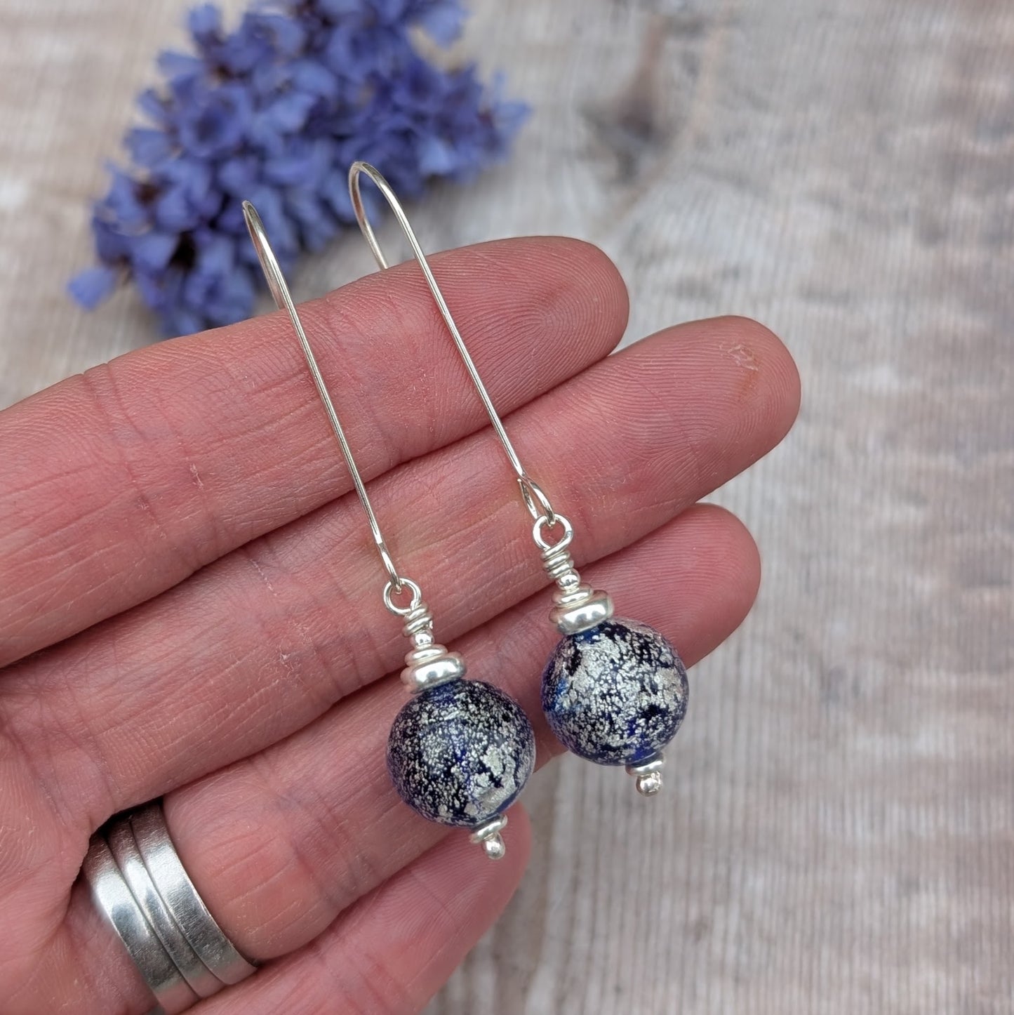 Silver blue venetian glass earrings displayed on a hand with dried flowers in the background.