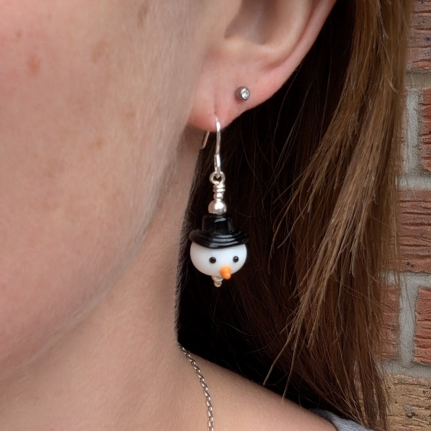 A person wearing one of the Silver Christmas Snowman Lampwork Glass Earrings, showing the handmade glass snowman bead with a black hat and orange nose hanging from a sterling silver hook.