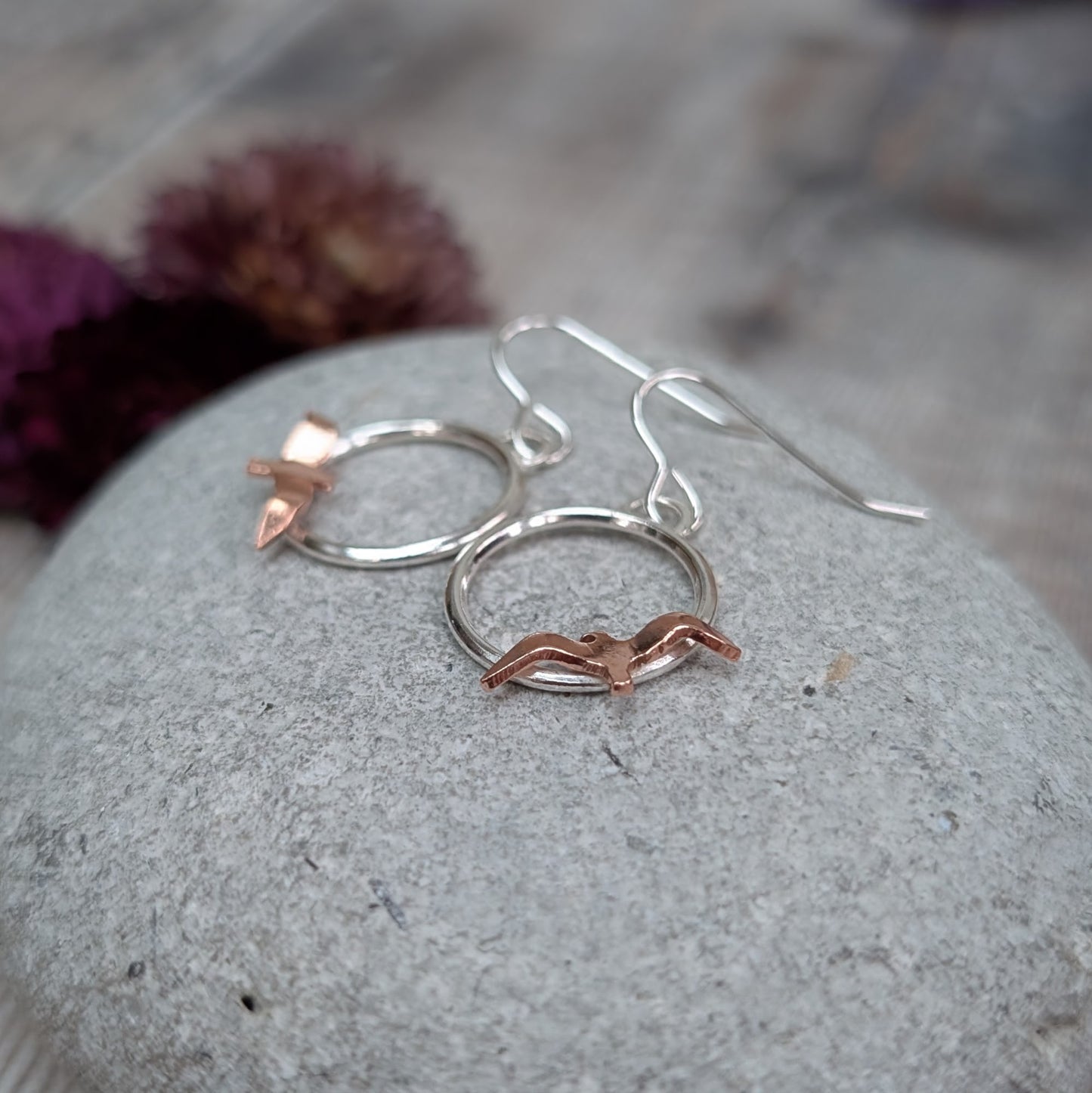 An angled close-up of Silver Circle Earrings with Copper Birds placed on a grey stone, highlighting the warm copper birds sitting on the lower edge of each silver circle.
