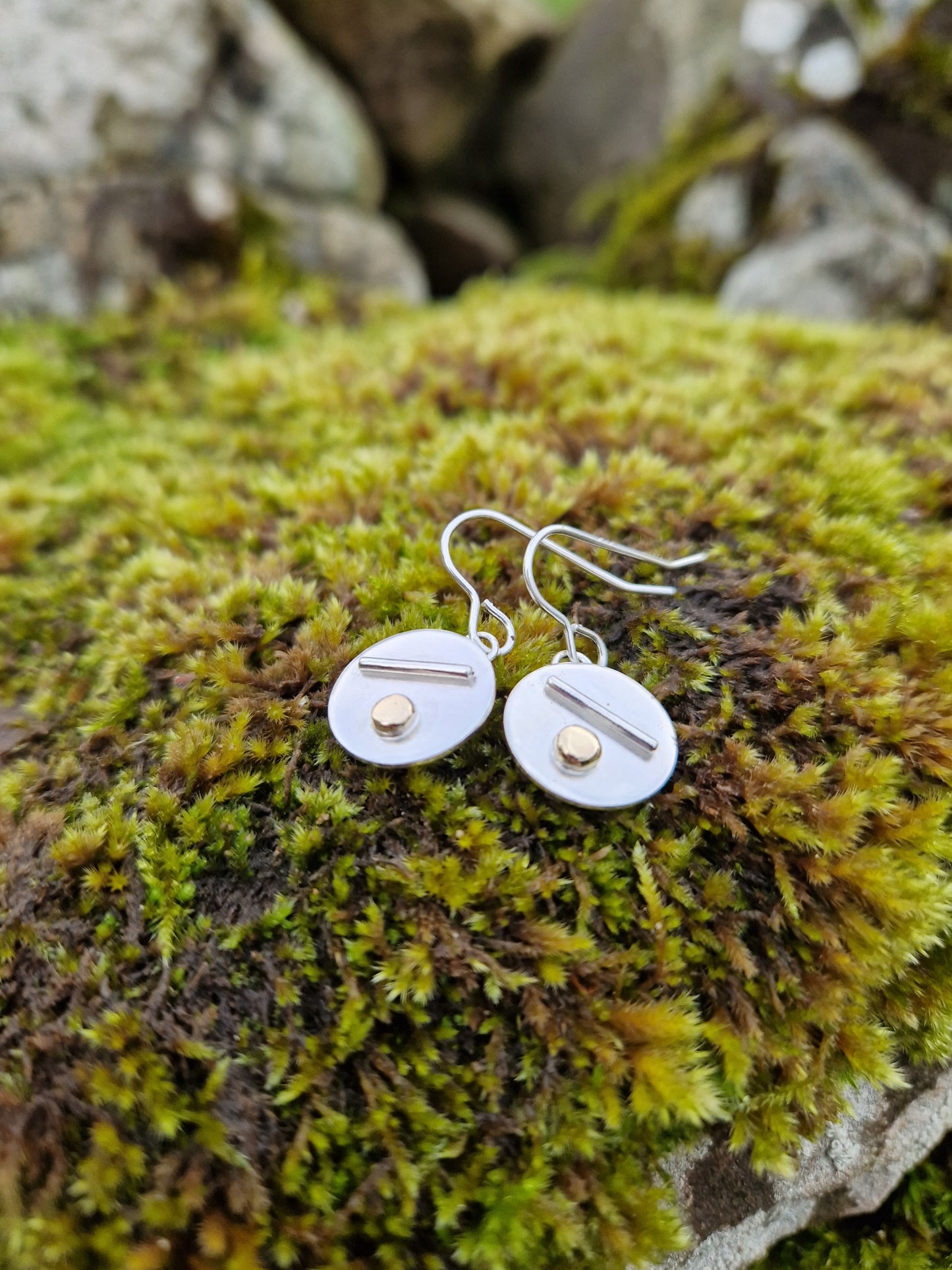 Each earring is a silver disc suspended from silver earring wire. On the disc is a raised silver line, off set to a gold raised round pebble shape underneath.