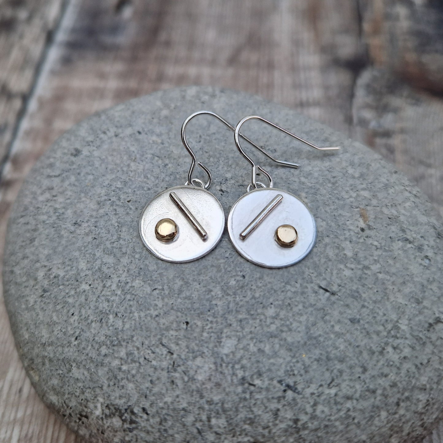 Each earring is a silver disc suspended from silver earring wire. On the disc is a raised silver line, off set to a gold raised round pebble shape underneath.