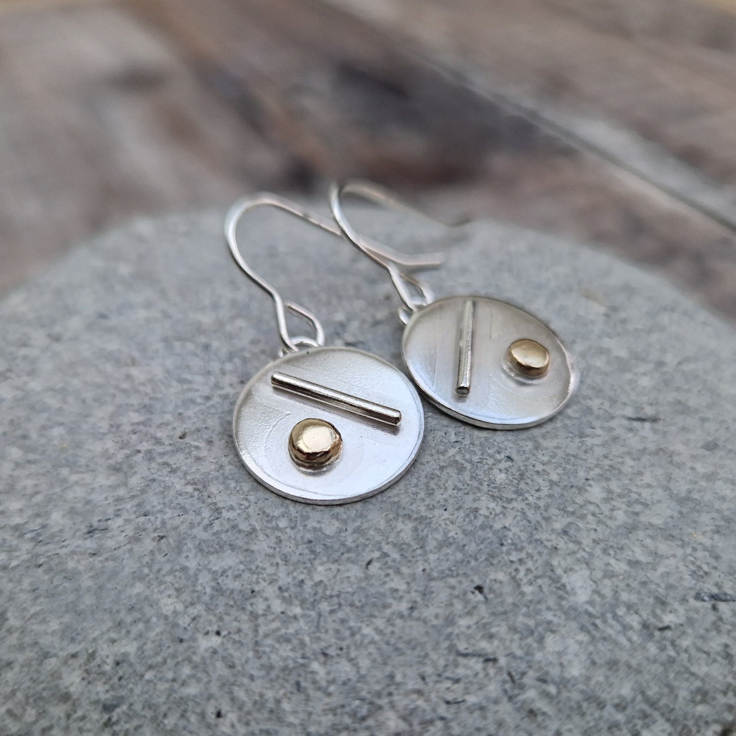Each earring is a silver disc suspended from silver earring wire. On the disc is a raised silver line, off set to a gold raised round pebble shape underneath.
