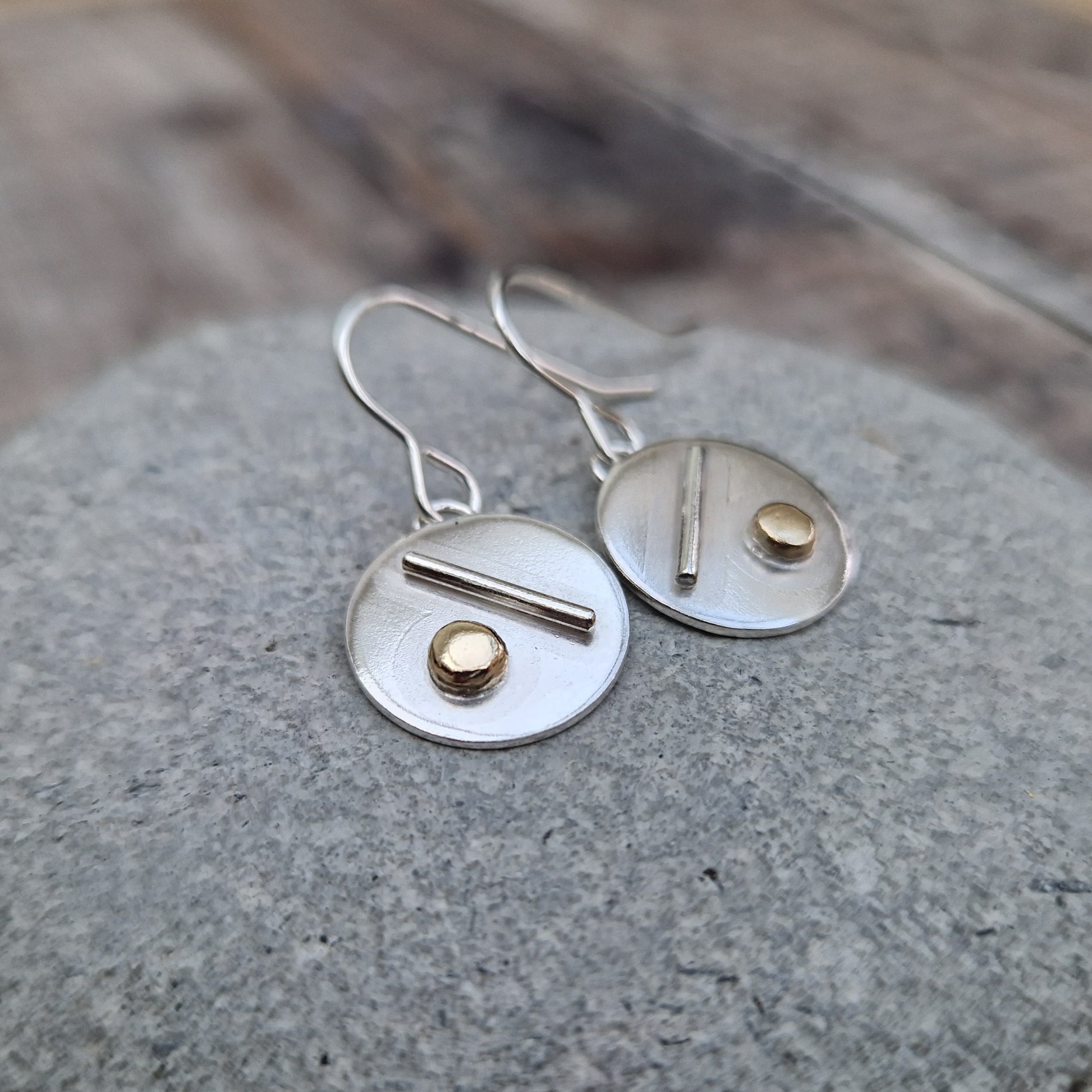 Each earring is a silver disc suspended from silver earring wire. On the disc is a raised silver line, off set to a gold raised round pebble shape underneath.