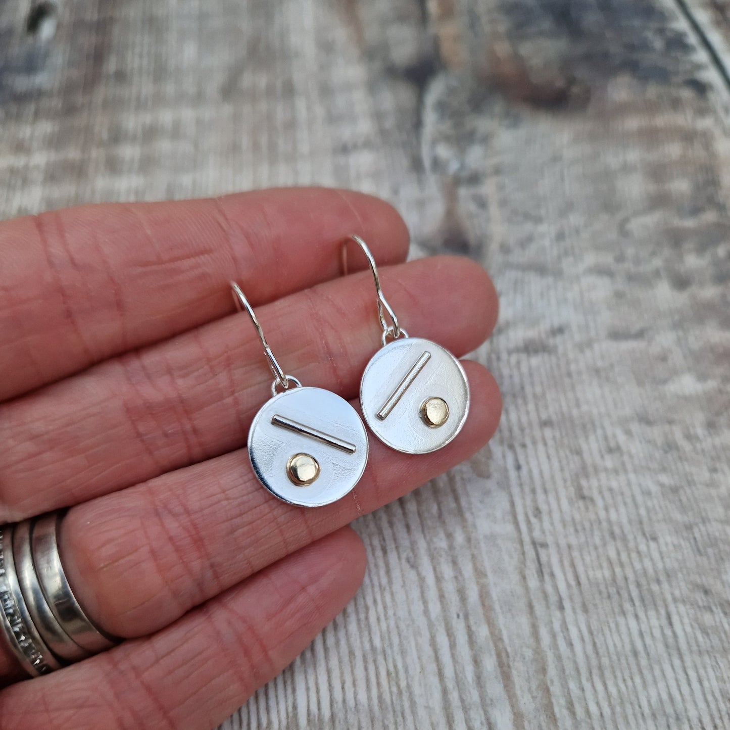 Each earring is a silver disc suspended from silver earring wire. On the disc is a raised silver line, off set to a gold raised round pebble shape underneath.