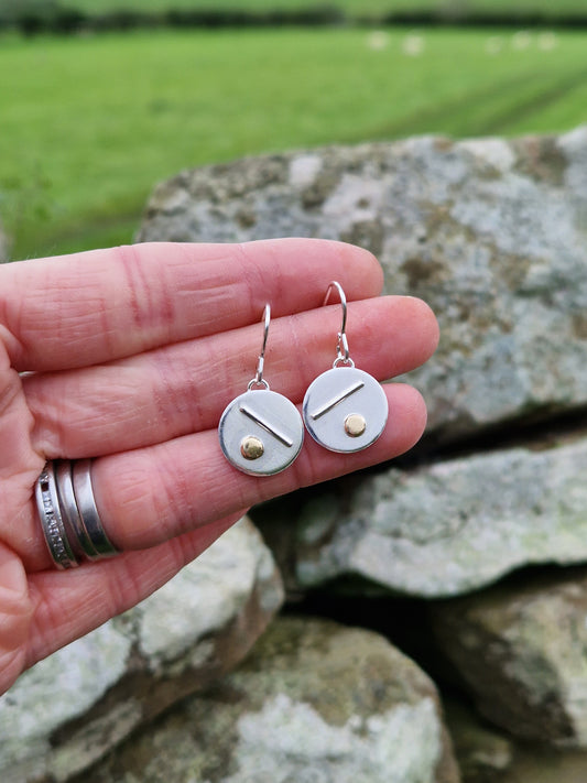 Each earring is a silver disc suspended from silver earring wire. On the disc is a raised silver line, off set to a gold raised round pebble shape underneath.