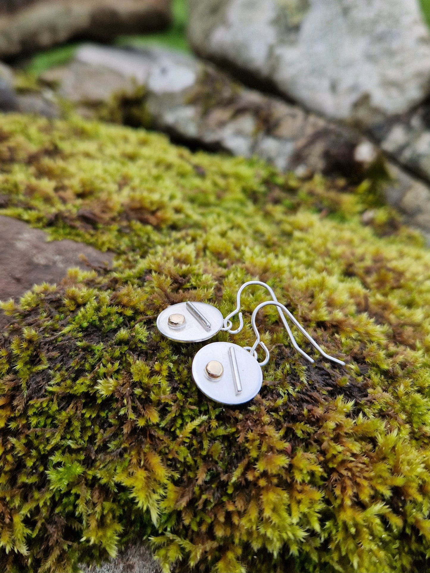 Each earring is a silver disc suspended from silver earring wire. On the disc is a raised silver line, off set to a gold raised round pebble shape underneath.