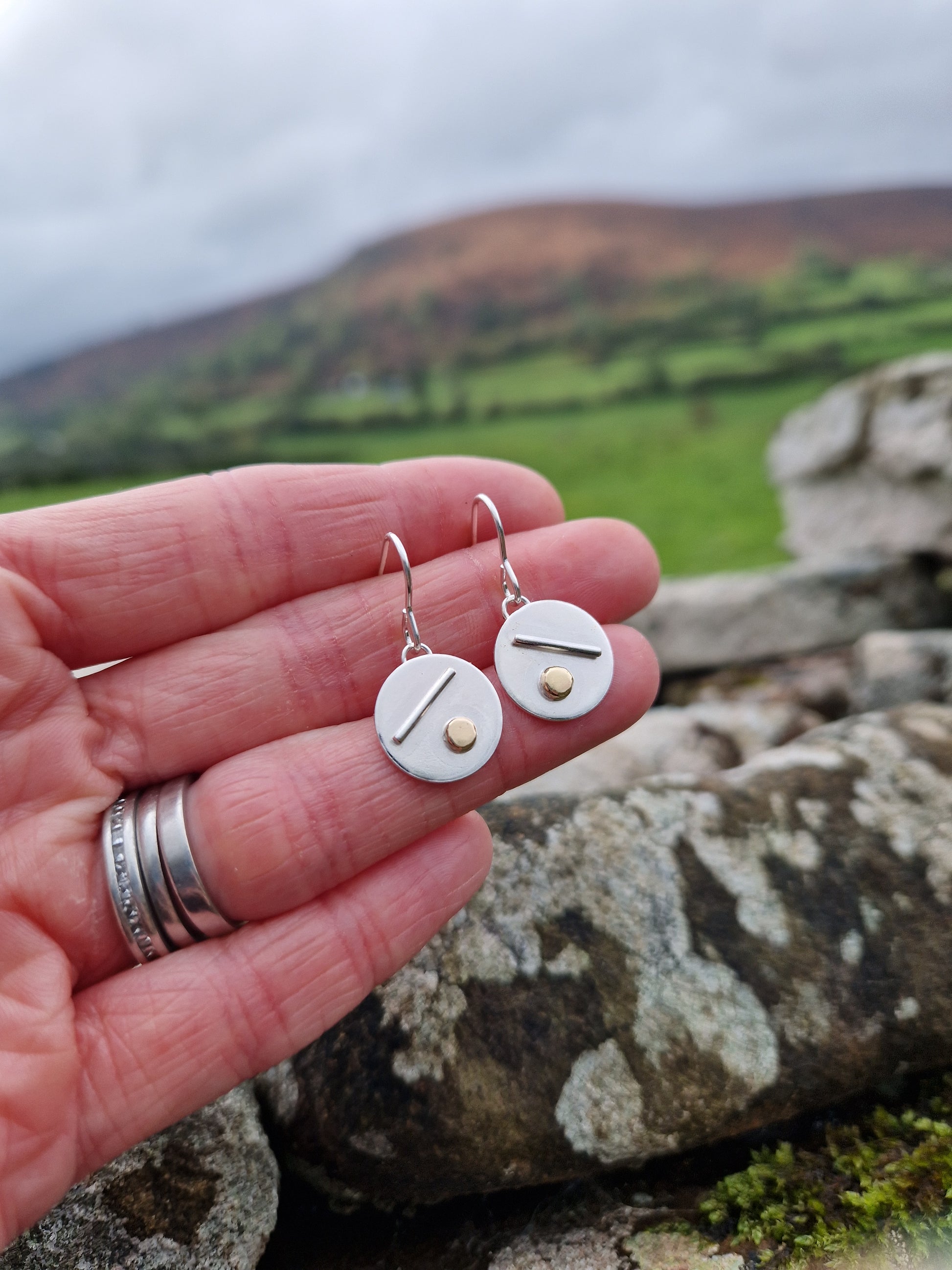 Each earring is a silver disc suspended from silver earring wire. On the disc is a raised silver line, off set to a gold raised round pebble shape underneath.