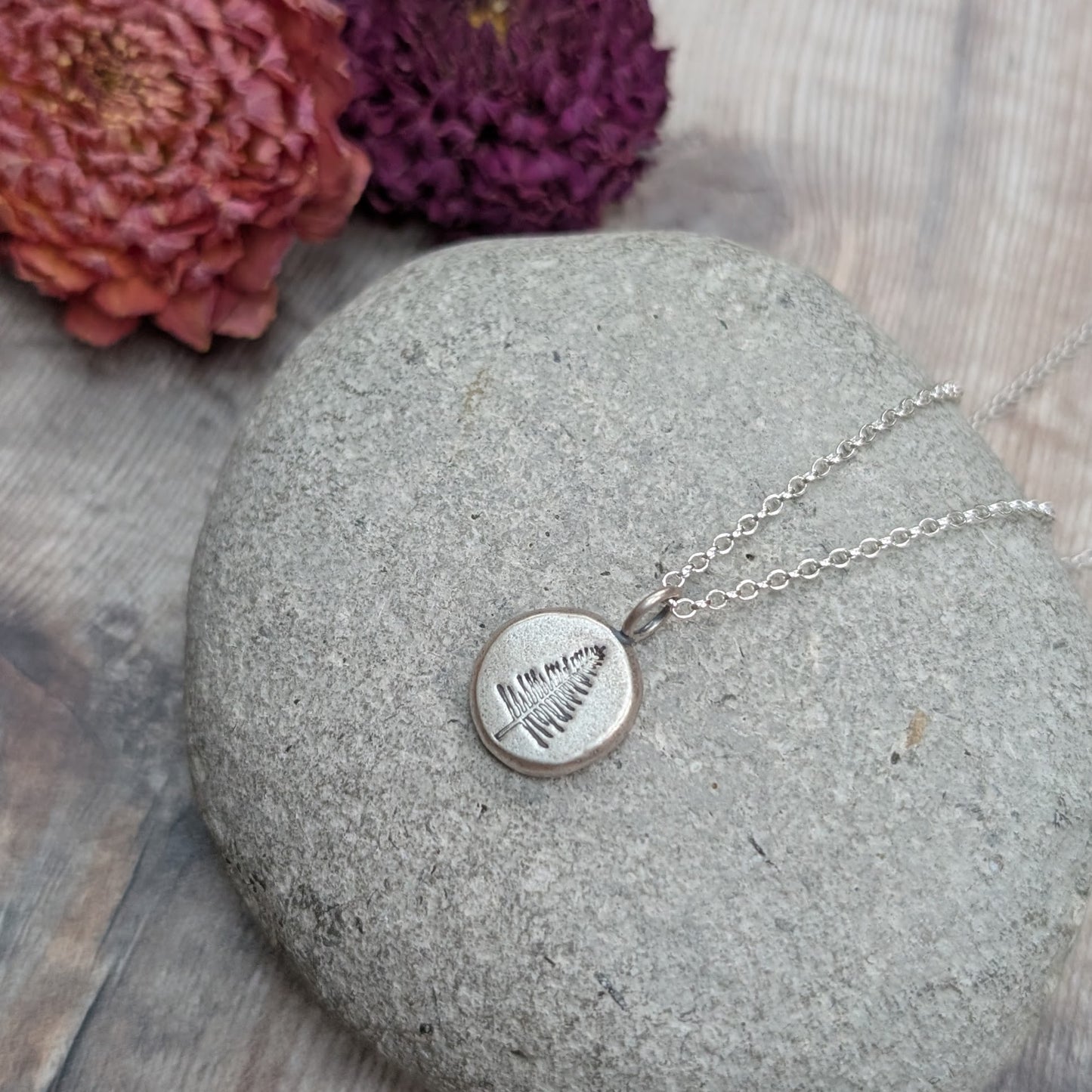 Silver evergreen tree pebble necklace resting on a smooth grey stone with dried flowers in the background.