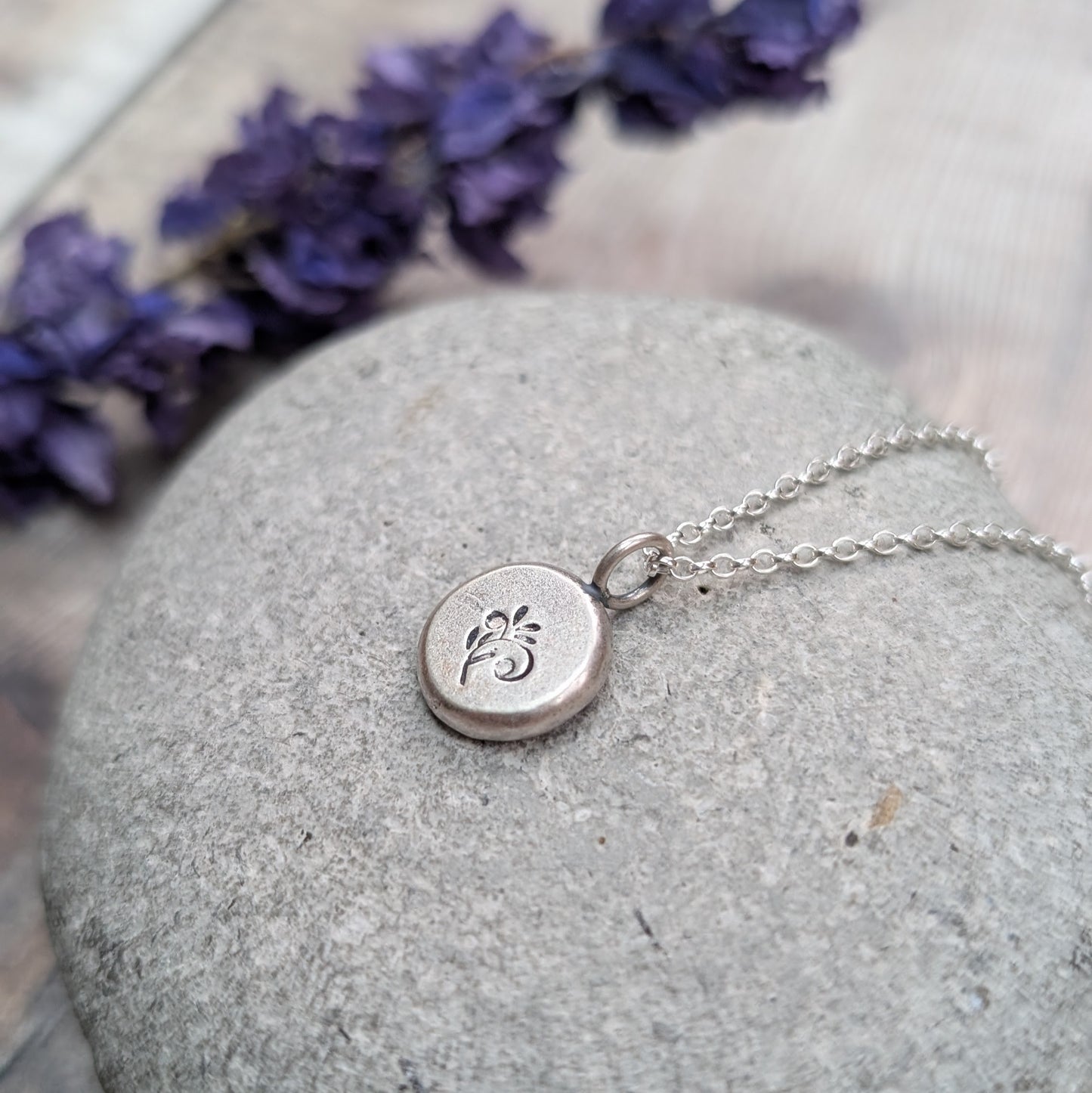Silver flower pebble necklace placed on a grey stone with a blurred sprig of purple flowers behind.