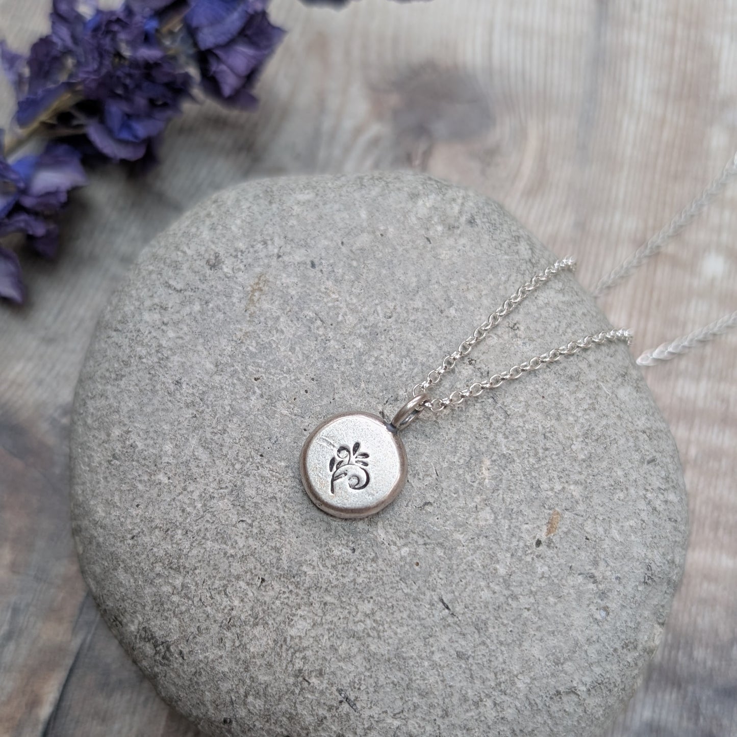 Silver flower pebble necklace resting on a smooth grey stone with purple flowers in the background.