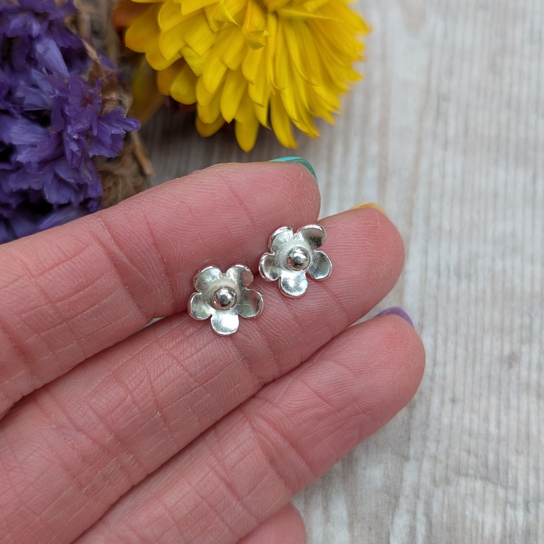Close-up of a hand holding a pair of handmade silver flower stud earrings with a blurred yellow and purple flower background.