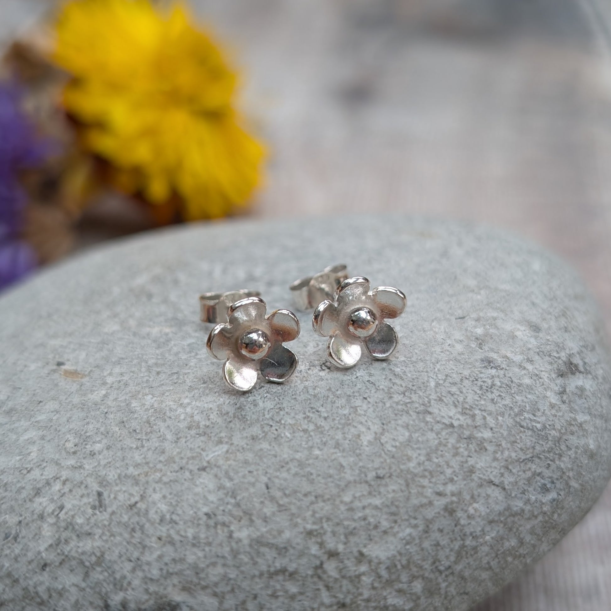 Silver flower stud earrings displayed on a smooth grey stone with blurred yellow and purple flowers in the background.