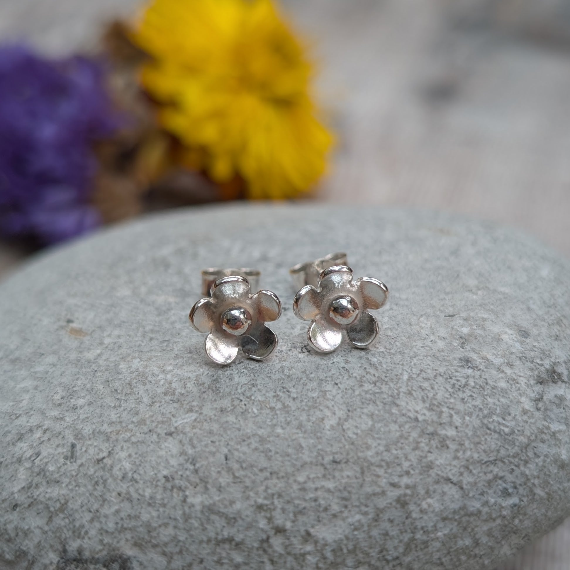 Pair of silver flower stud earrings photographed on a grey stone surface with a colourful floral backdrop.