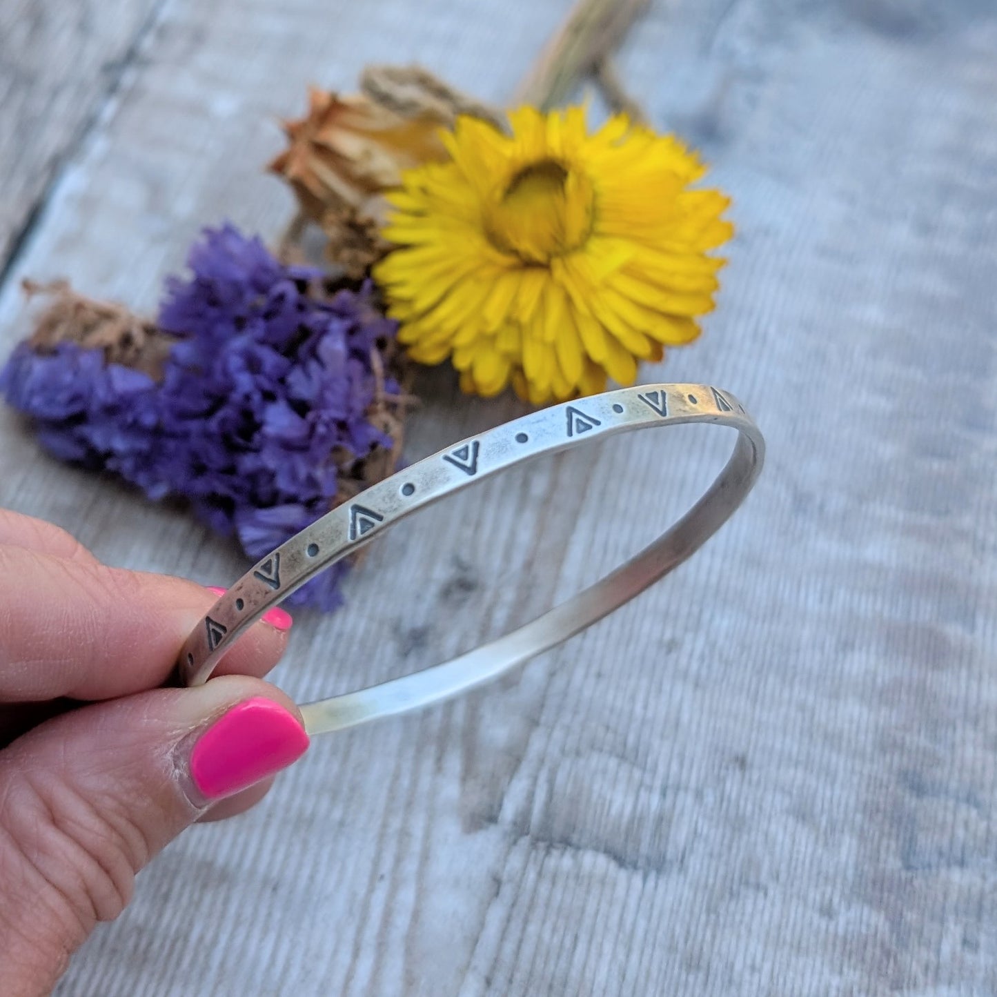 A hand holding the silver geometric patterned bangle in front of dried yellow and purple flowers on a wooden surface.