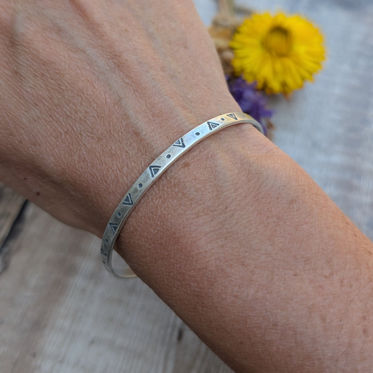 A silver geometric patterned bangle worn on a wrist, showing the hand-stamped triangle and dot design.