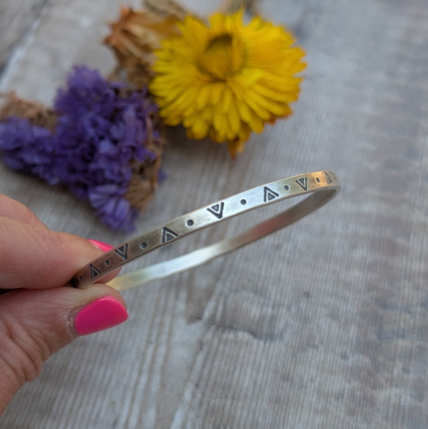 A hand holding the silver geometric patterned bangle in front of dried yellow and purple flowers on a wooden surface.