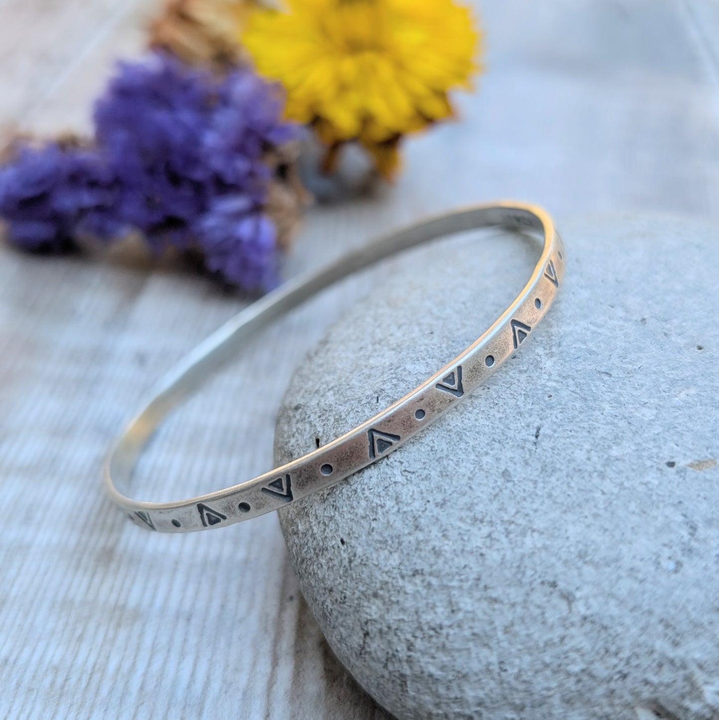Close-up of the silver geometric patterned bangle resting on a stone with yellow and purple flowers in the background.
