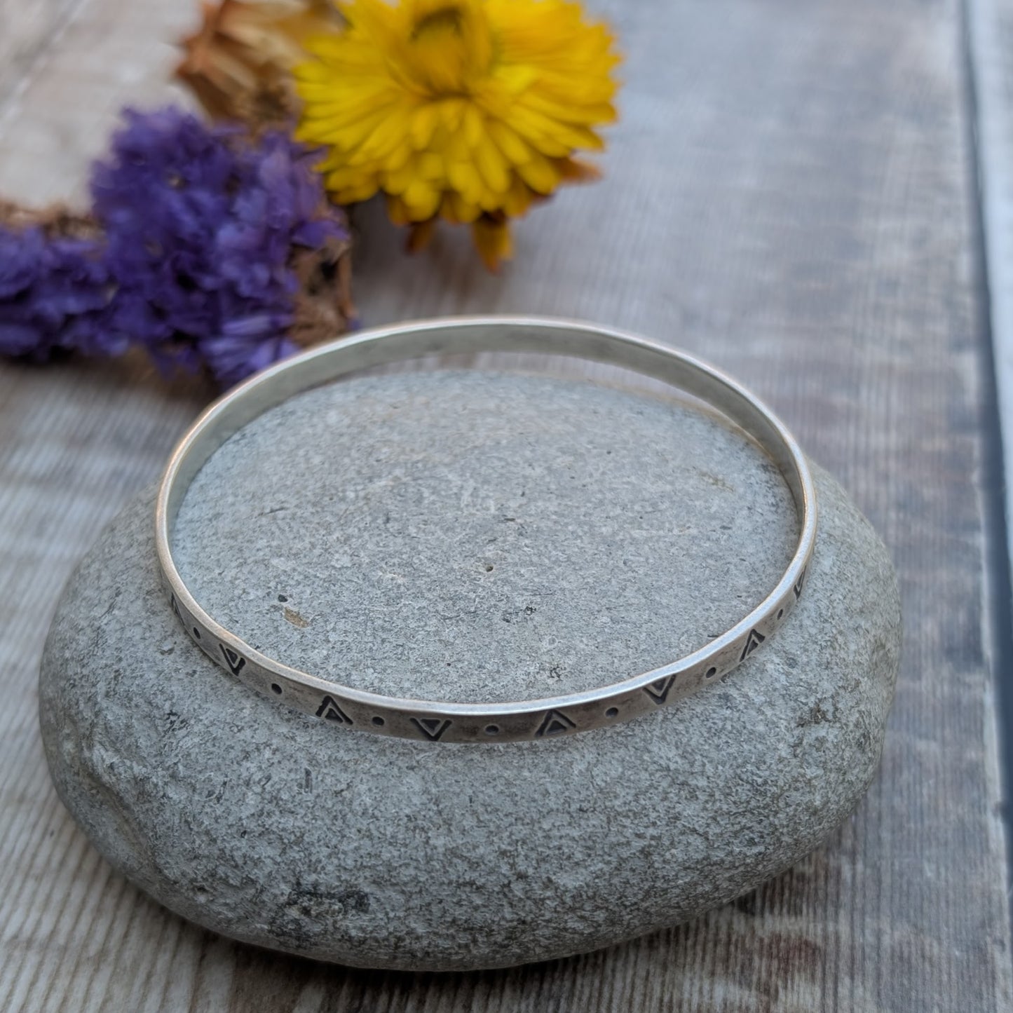 Silver geometric patterned bangle displayed flat on a smooth stone, showing the full circular shape and stamped detail.