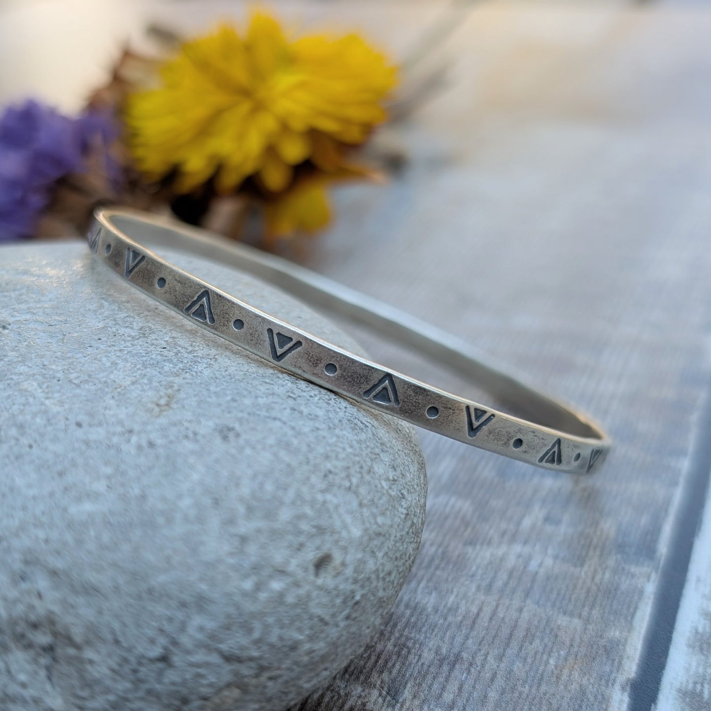 Close-up of the silver geometric patterned bangle resting on a stone with yellow and purple flowers in the background.